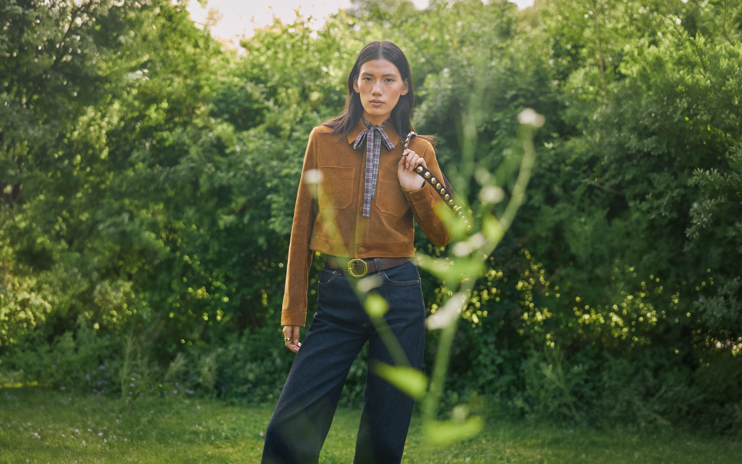 A woman wearing a tan suede jacket and dark denim jeans stands in a lush green garden.