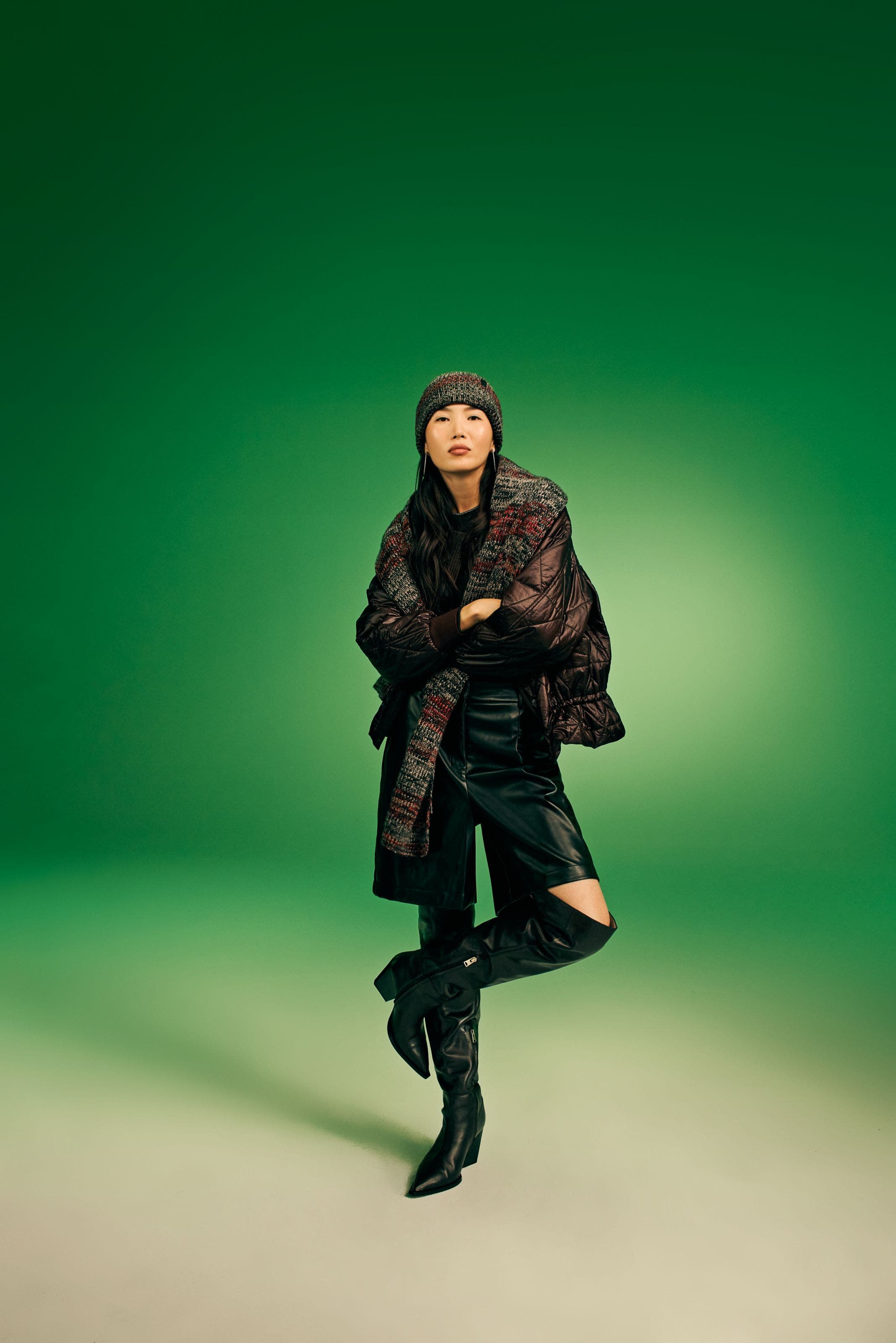 Model wearing a dark quilted puffer jacket, multicolor knit scarf and beanie, black leather pants, and black knee-high boots.