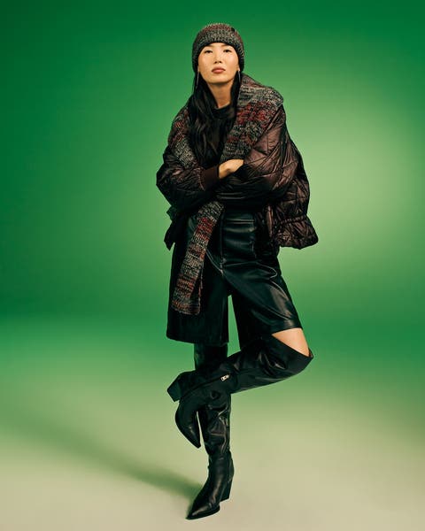 Model wearing a dark quilted puffer jacket, multicolor knit scarf and beanie, black leather pants, and black knee-high boots.