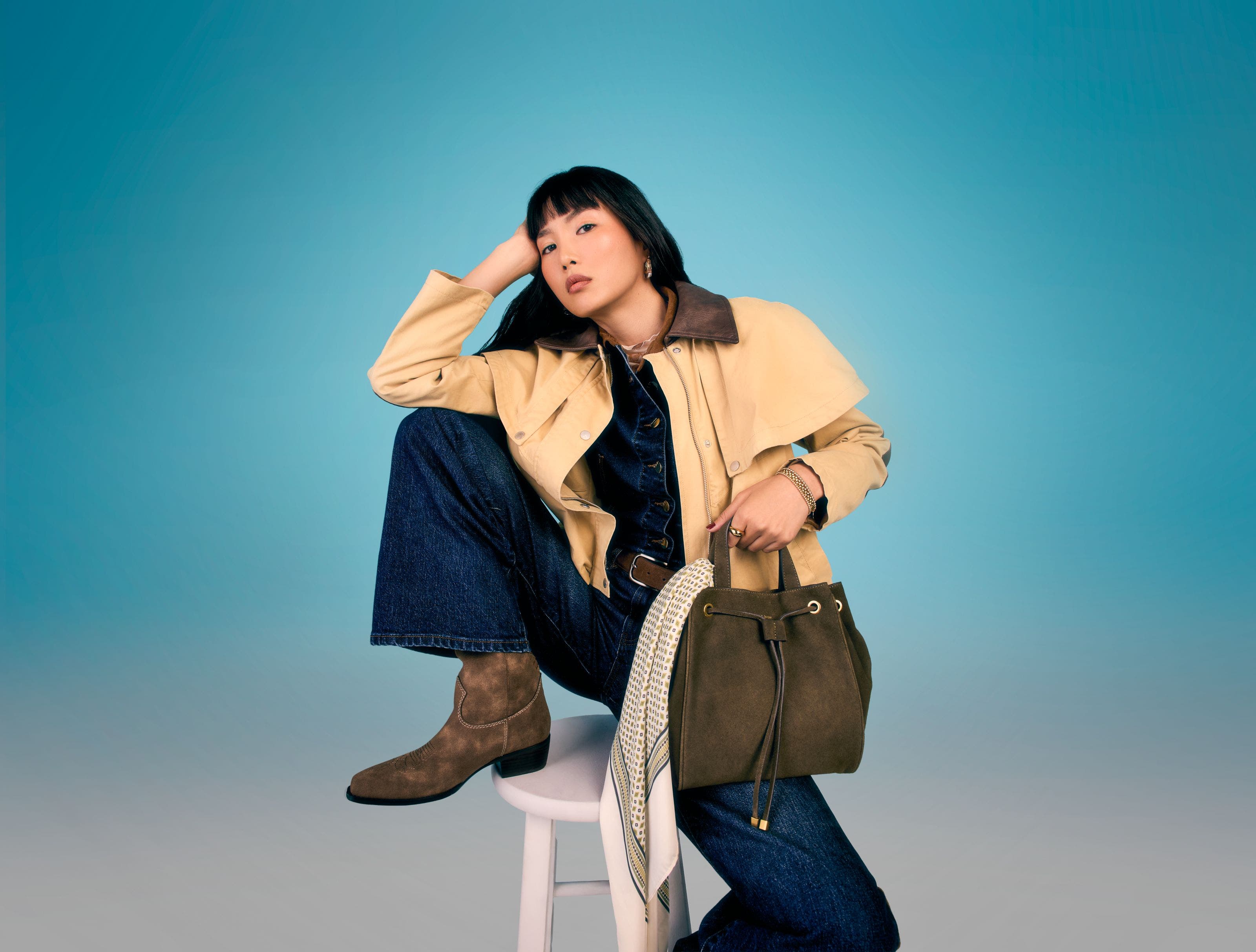 A woman wearing a blue denim jumpsuit, suede ankle boots and carrying a suede bucket bag.