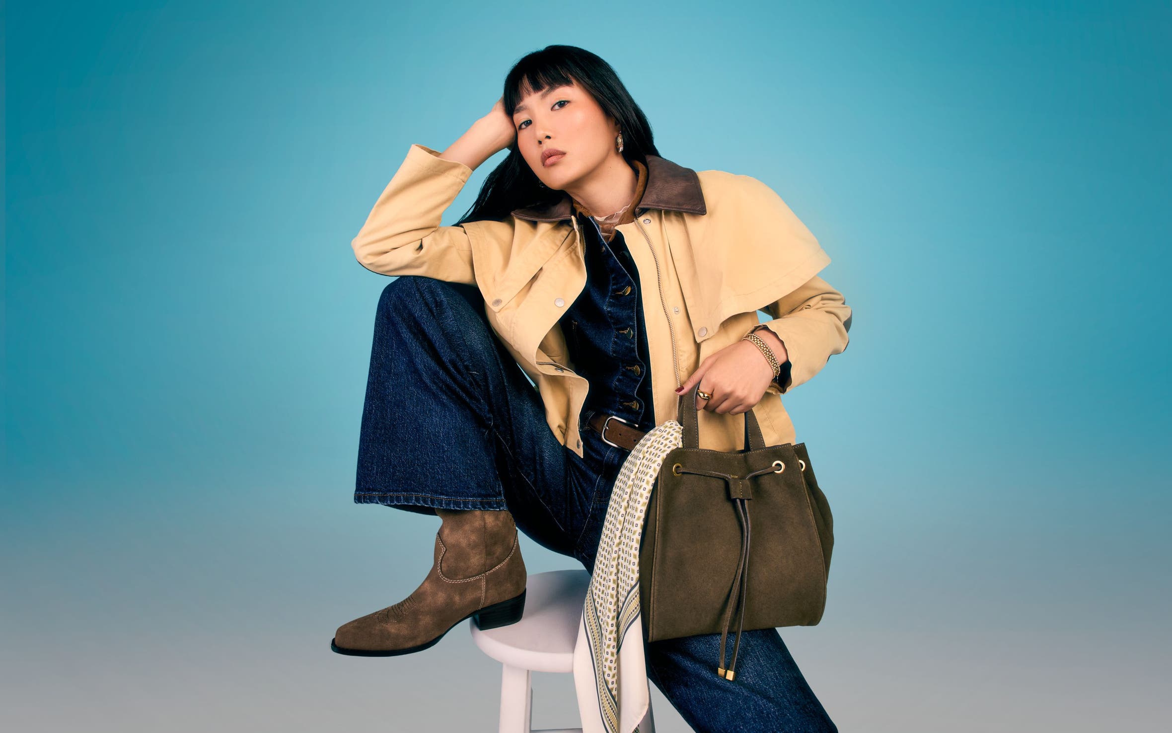 A woman wearing a blue denim jumpsuit, suede ankle boots and carrying a suede bucket bag.