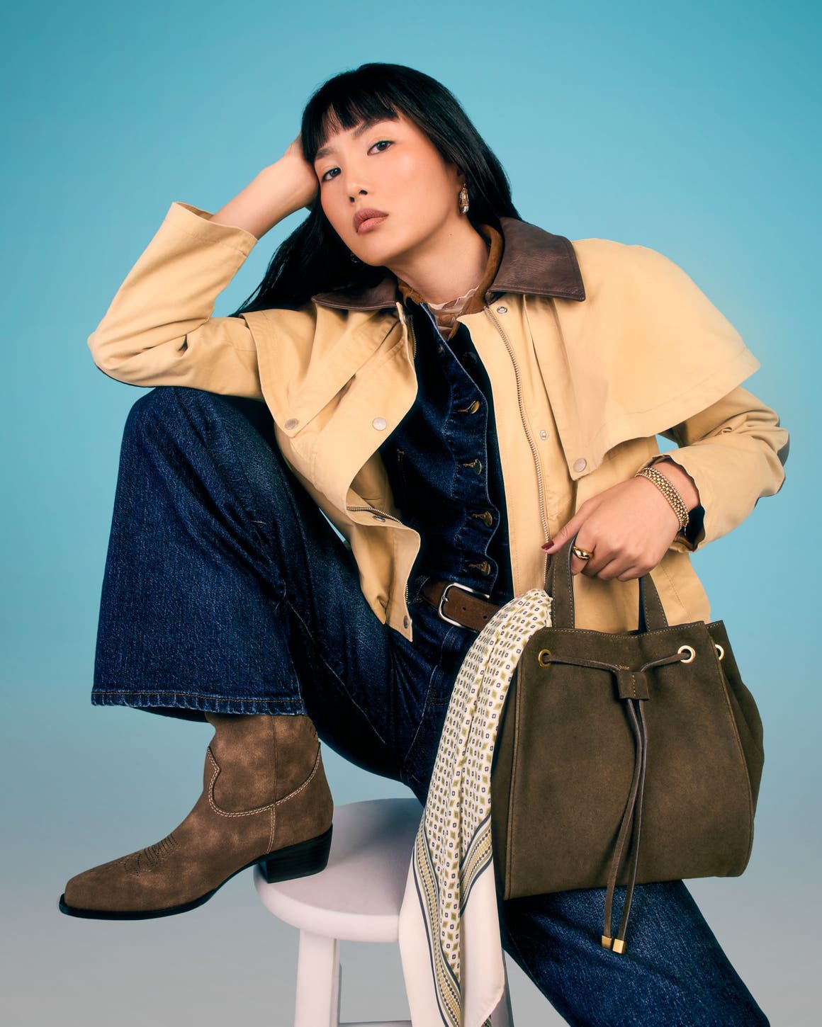 A woman wearing a blue denim jumpsuit, suede ankle boots and carrying a suede bucket bag.