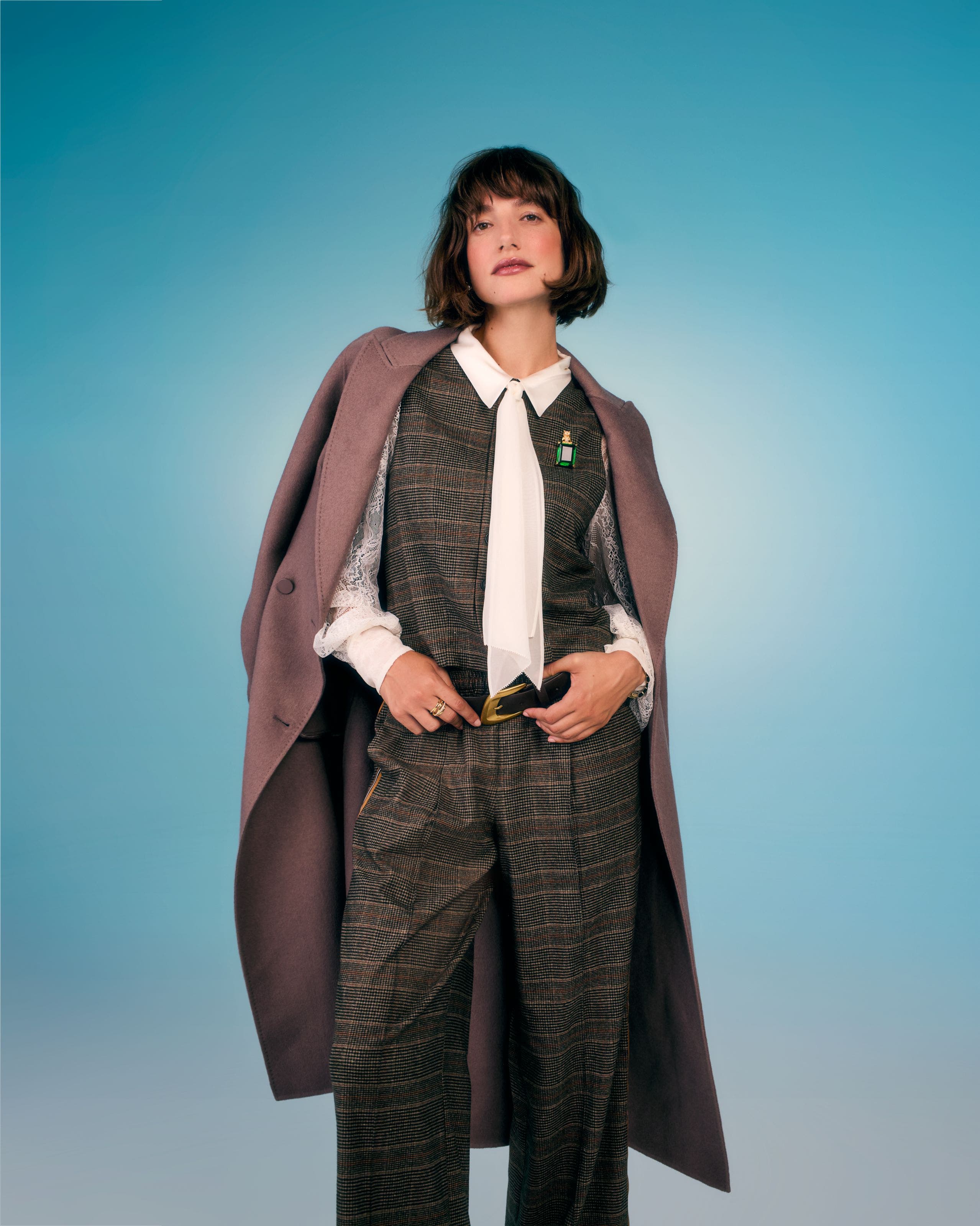 A woman wearing a plaid vest and trousers suit set with a white blouse and draped coat.