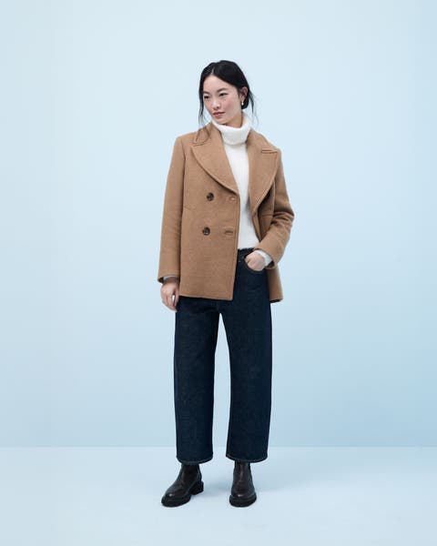 Woman wearing a white turtleneck sweater, camel-colored pea coat, jeans and boots.