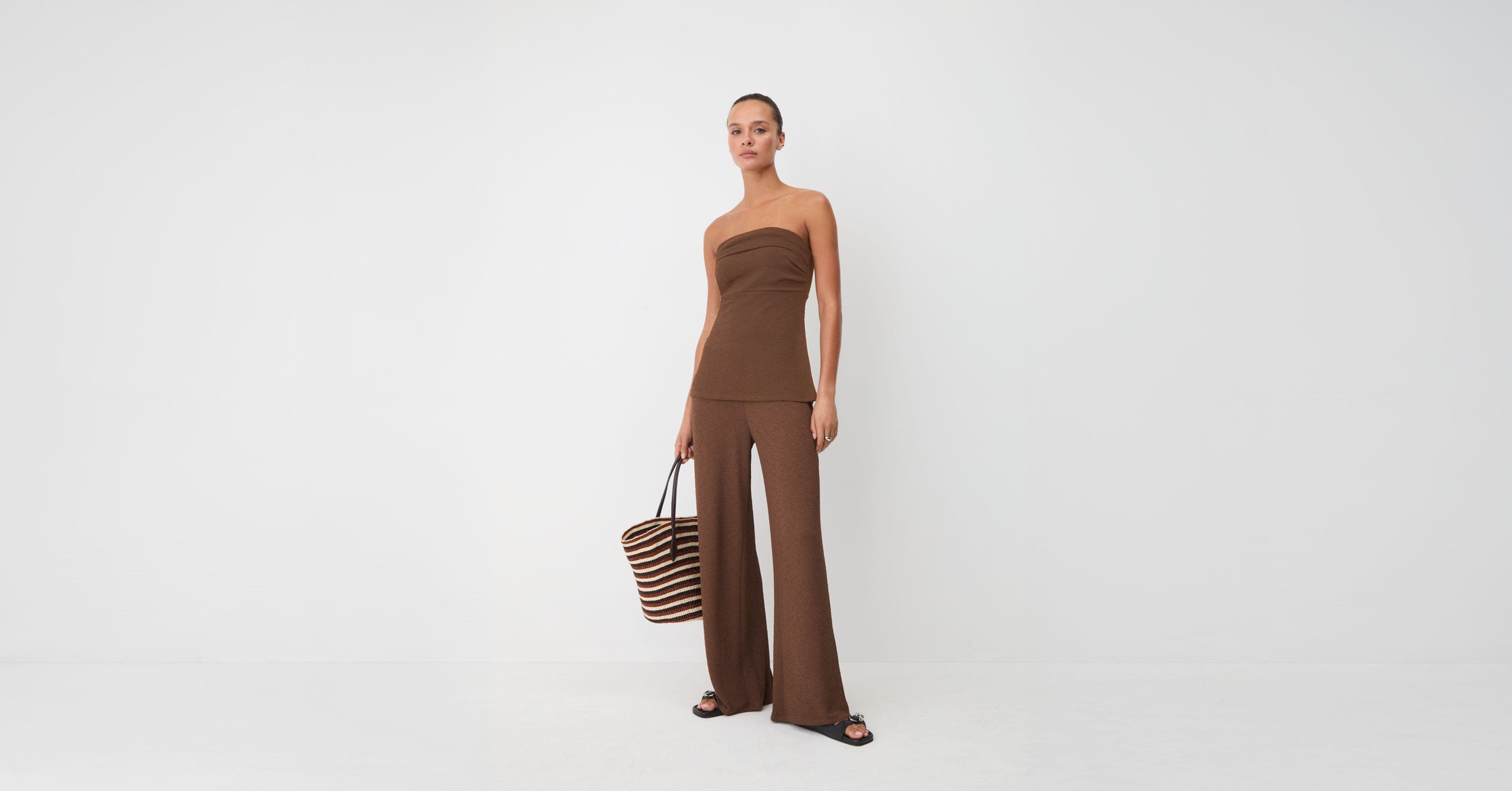 Woman wearing a matching brown strapless top and wide legged pants carrying a tote.