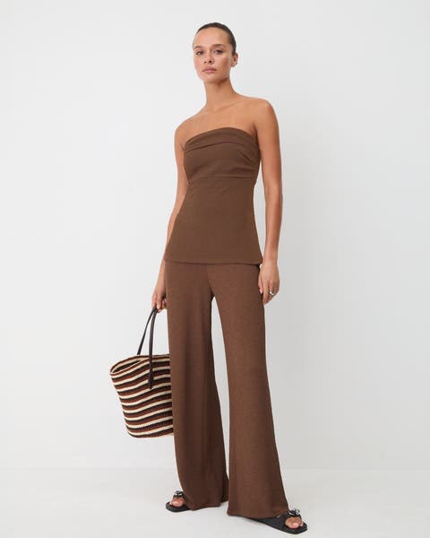 Woman wearing a matching brown strapless top and wide legged pants carrying a tote.