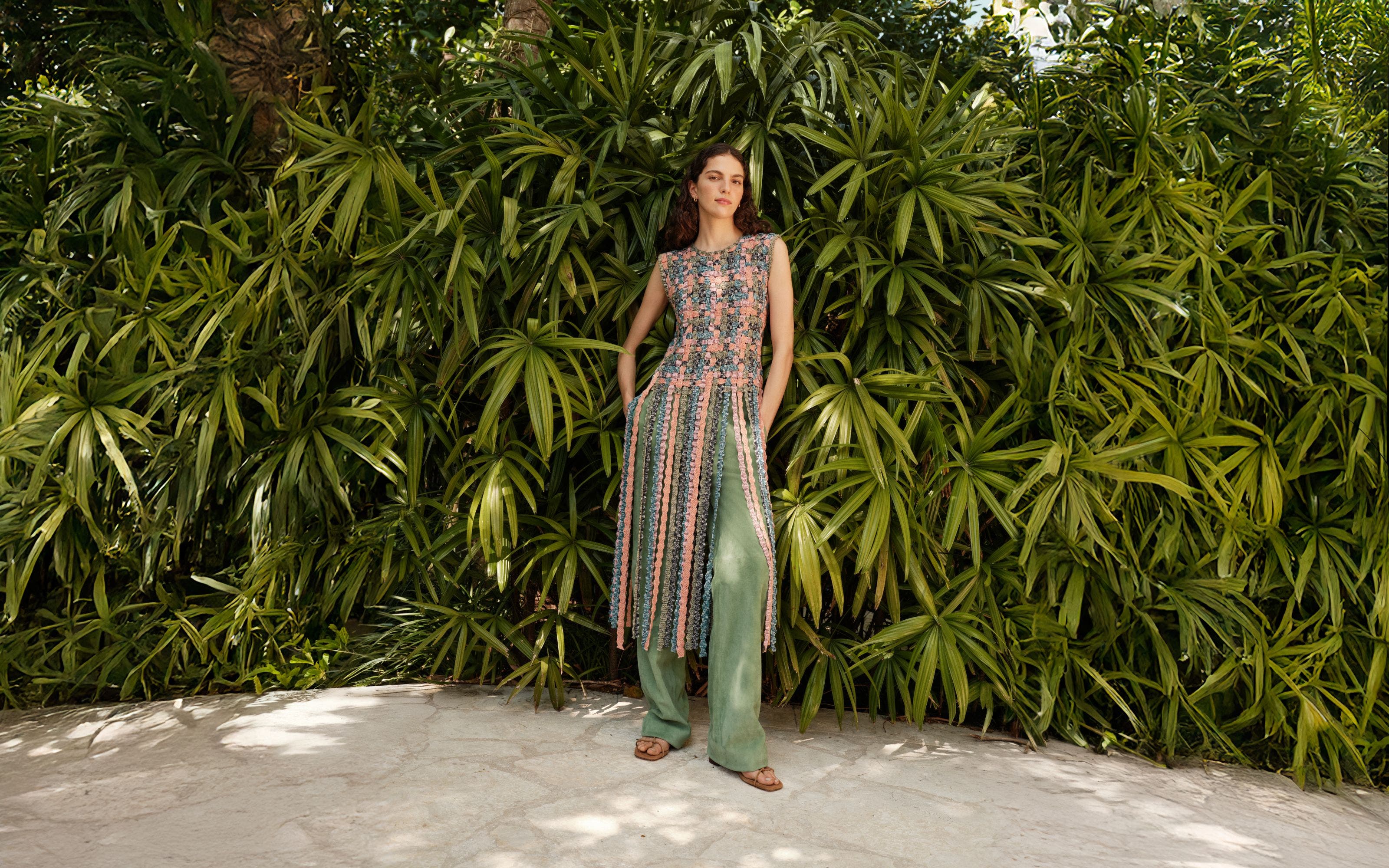 A woman wearing a knitted top with fringe, green pants and sandals.
