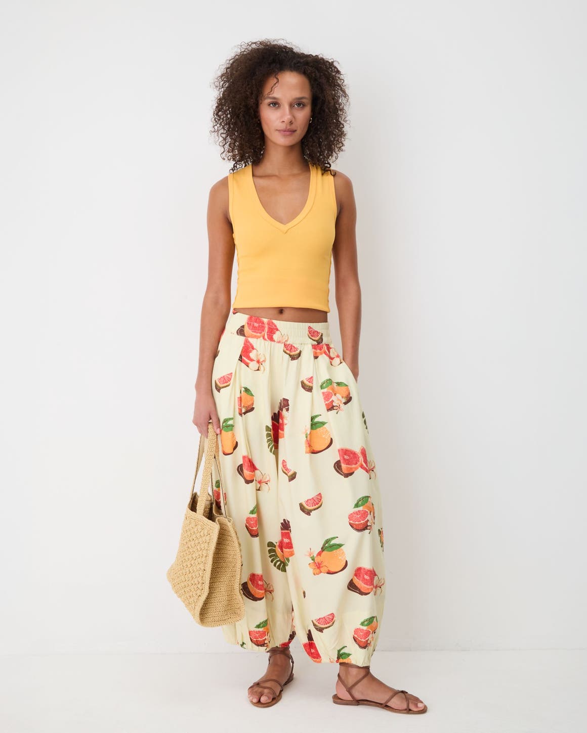 A woman wearing an orange Free People crop top with fruit-printed pants, sandals and a woven tote bag. 