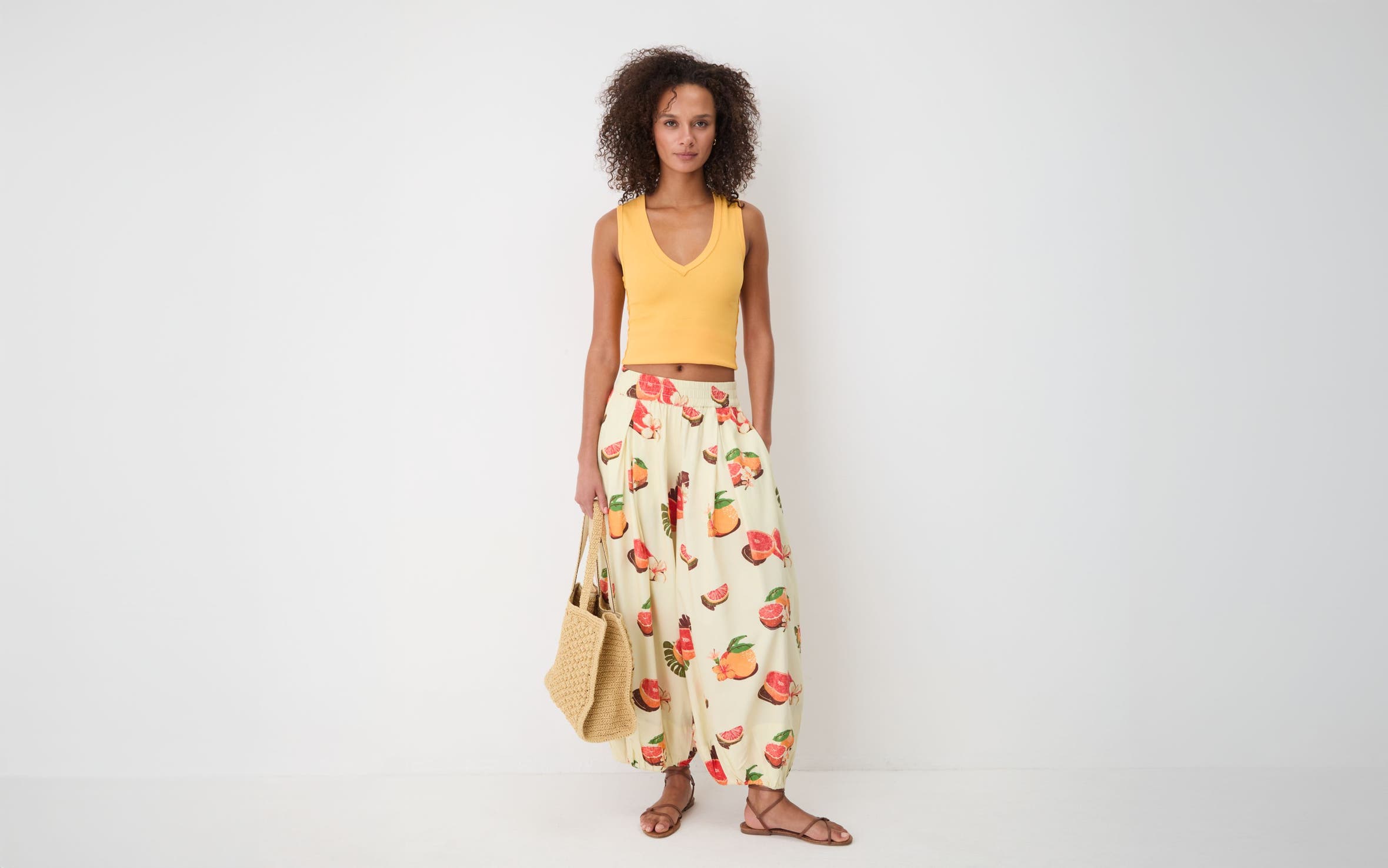 A woman wearing an orange Free People crop top with fruit-printed pants, sandals and a woven tote bag. 