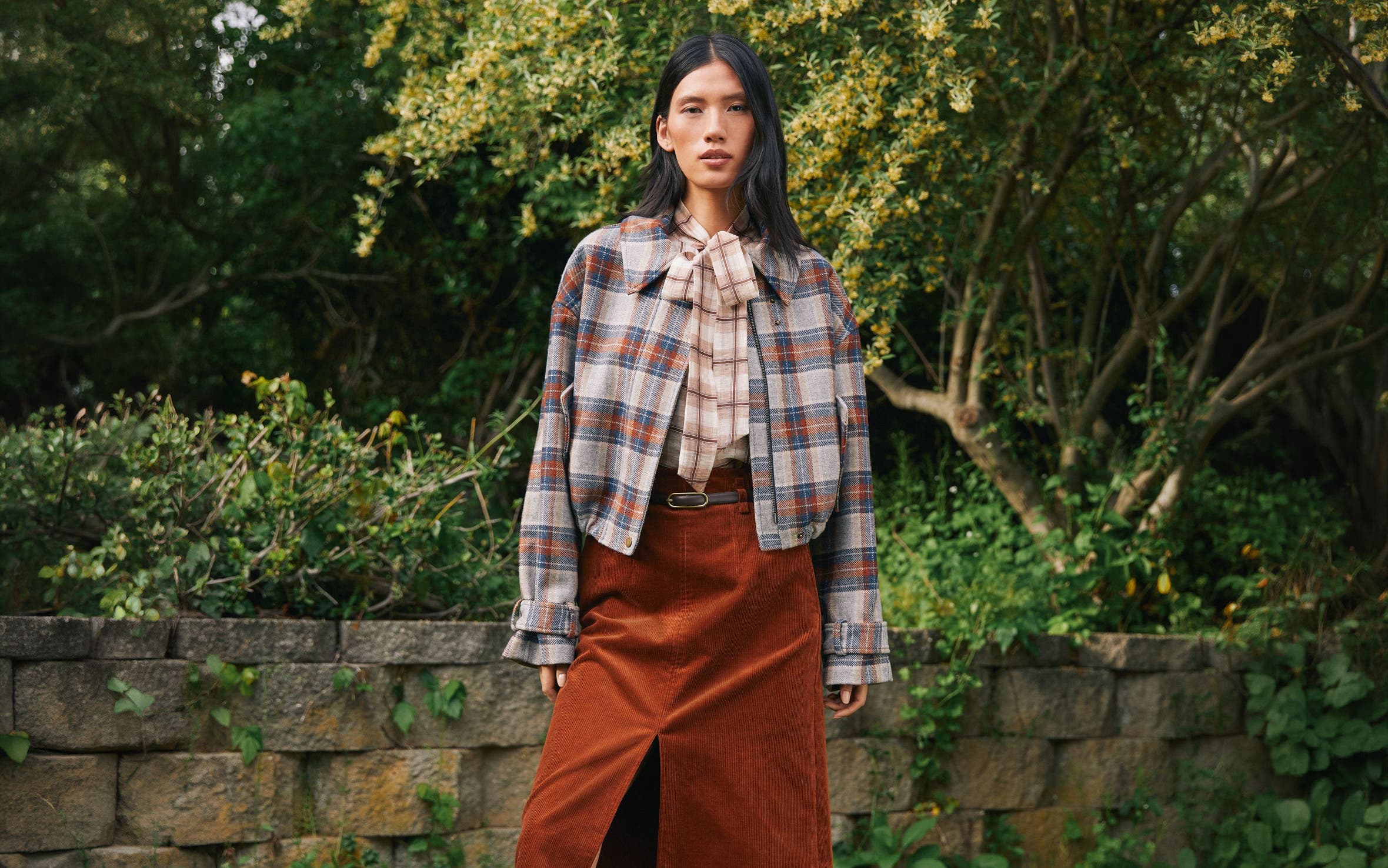 A woman wearing a plaid shirt, plaid jacket, and brown midi skirt.