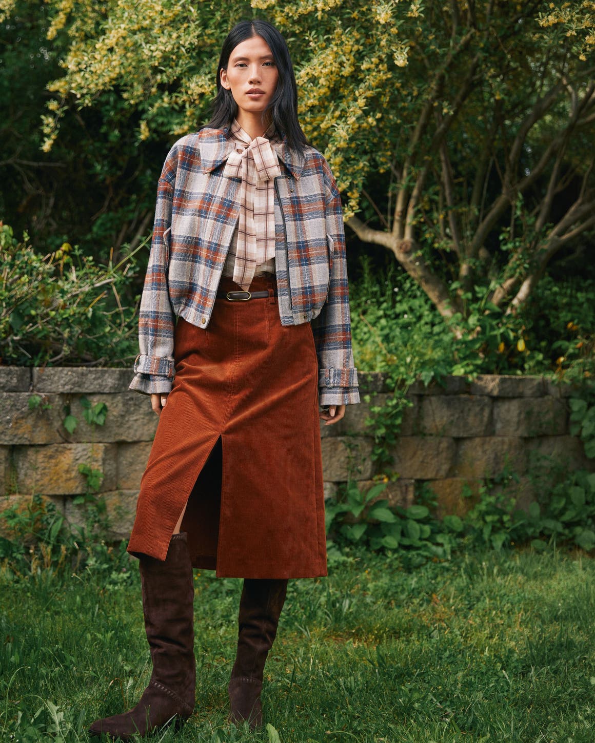 A woman wearing a plaid shirt, plaid jacket, and brown midi skirt.