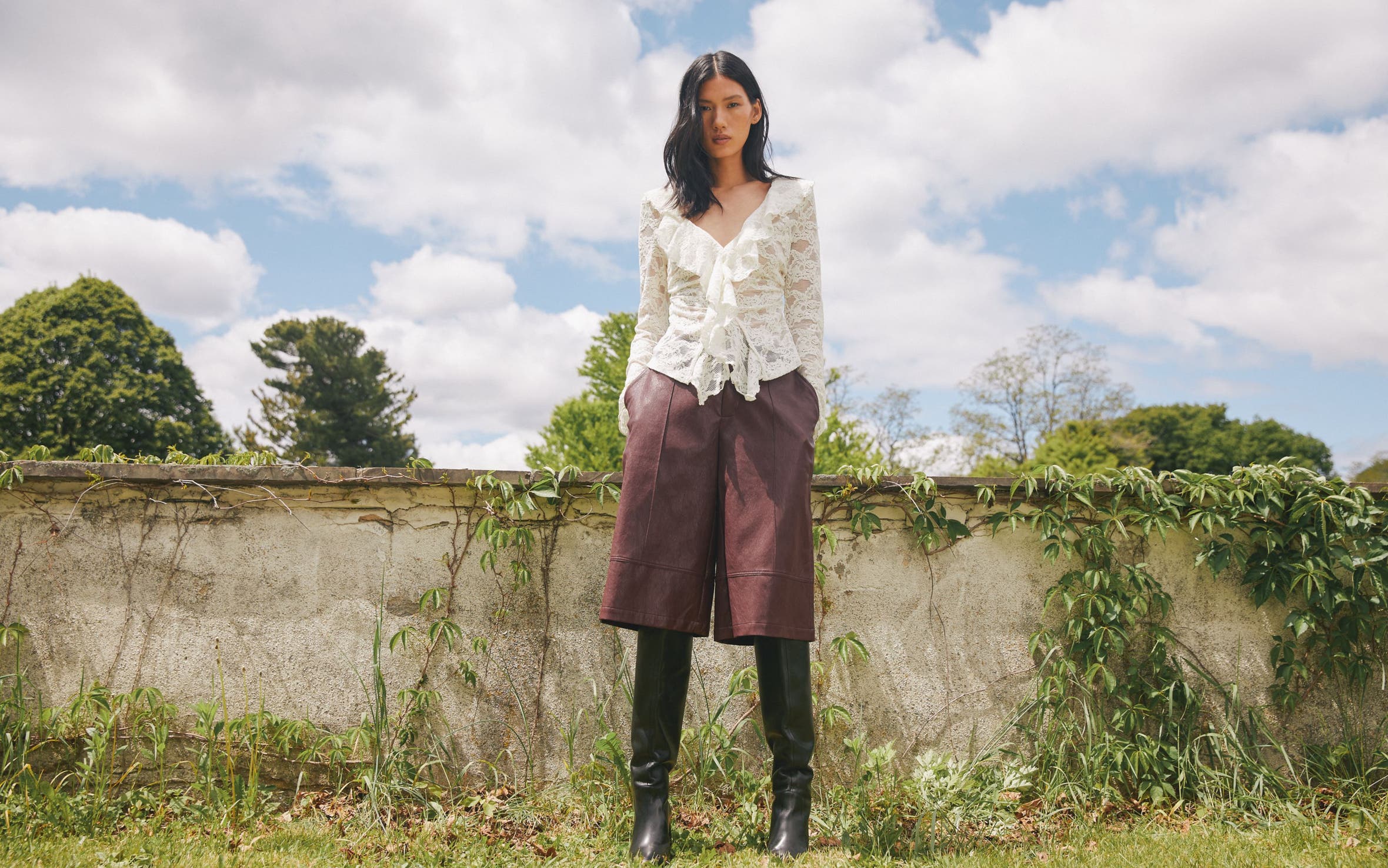A woman wearing a white lace long-sleeve blouse with ruffle details, brown knee-length culottes, and tall black boots.