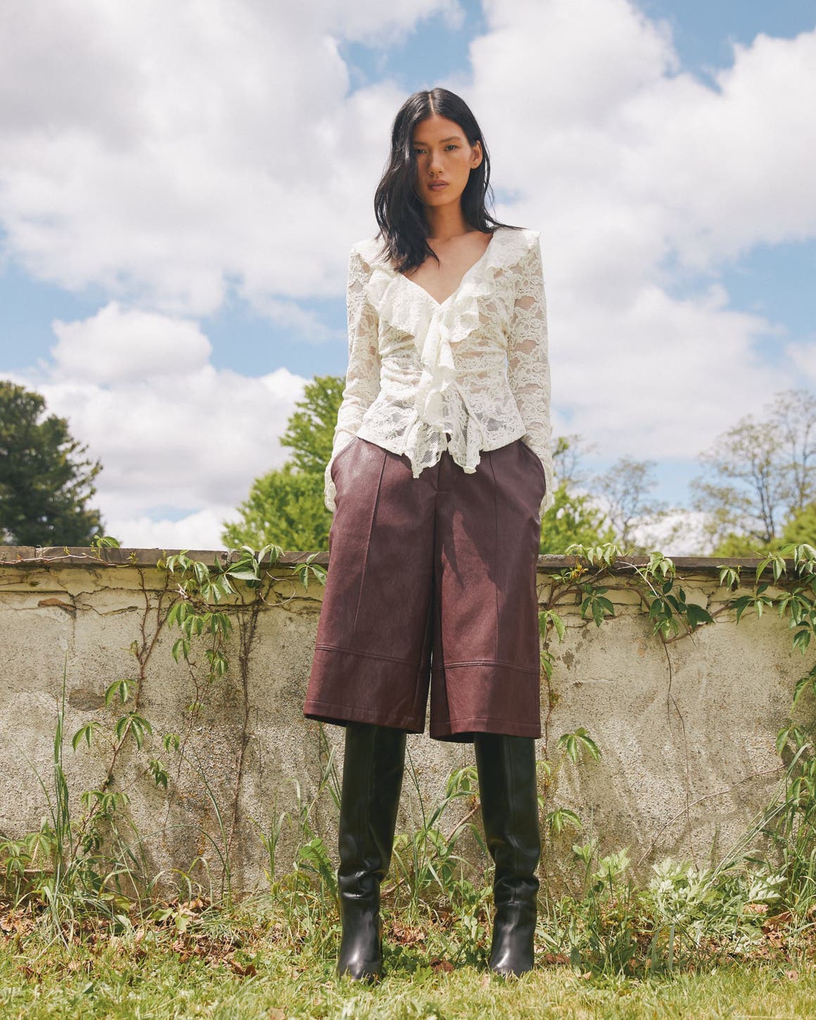 A woman wearing a white lace long-sleeve blouse with ruffle details, brown knee-length culottes, and tall black boots.