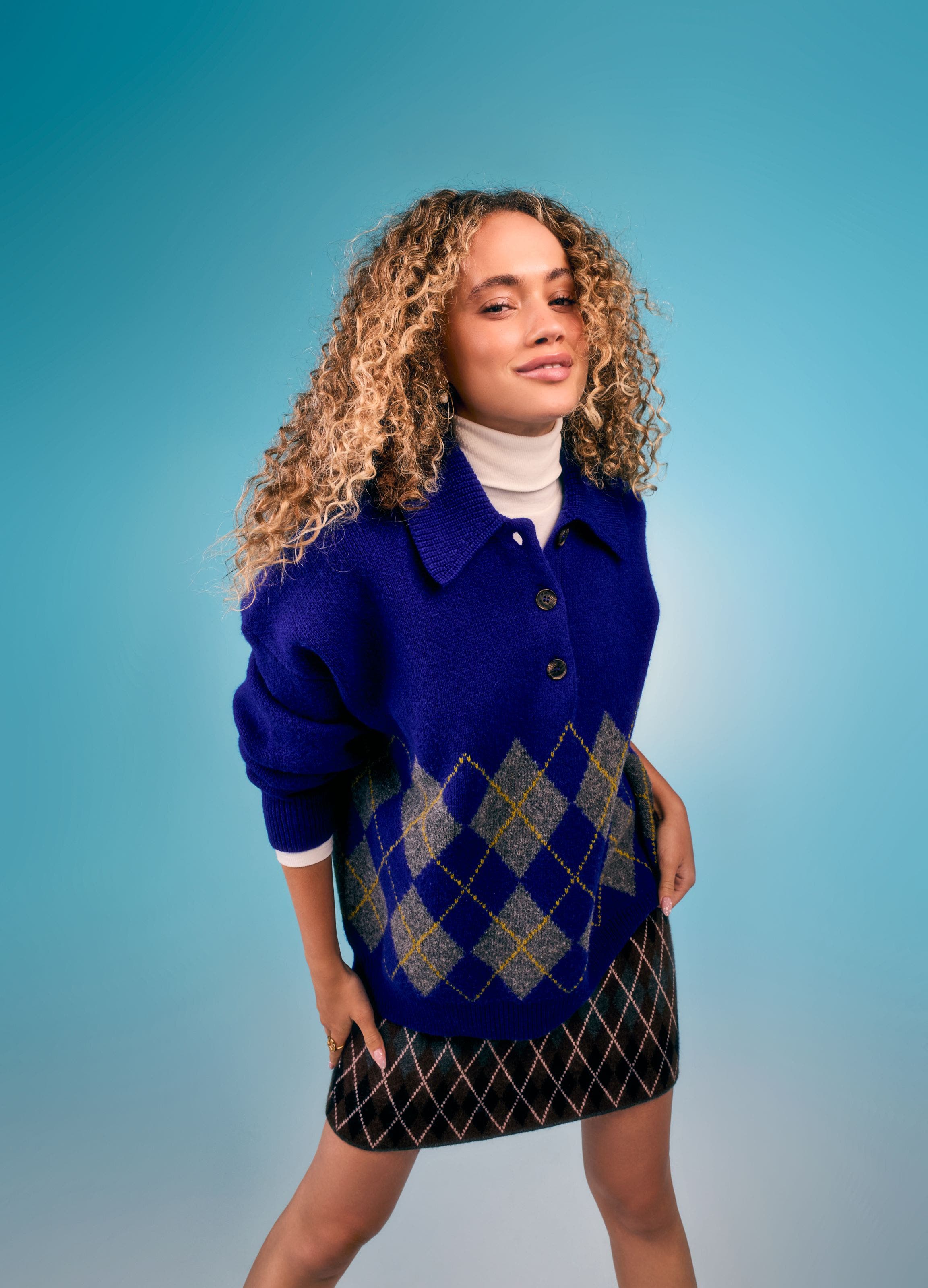 Woman wearing a royal blue buttoned sweater with gray-green argyle pattern, layered over a white turtleneck and paired with a dark plaid mini skirt.