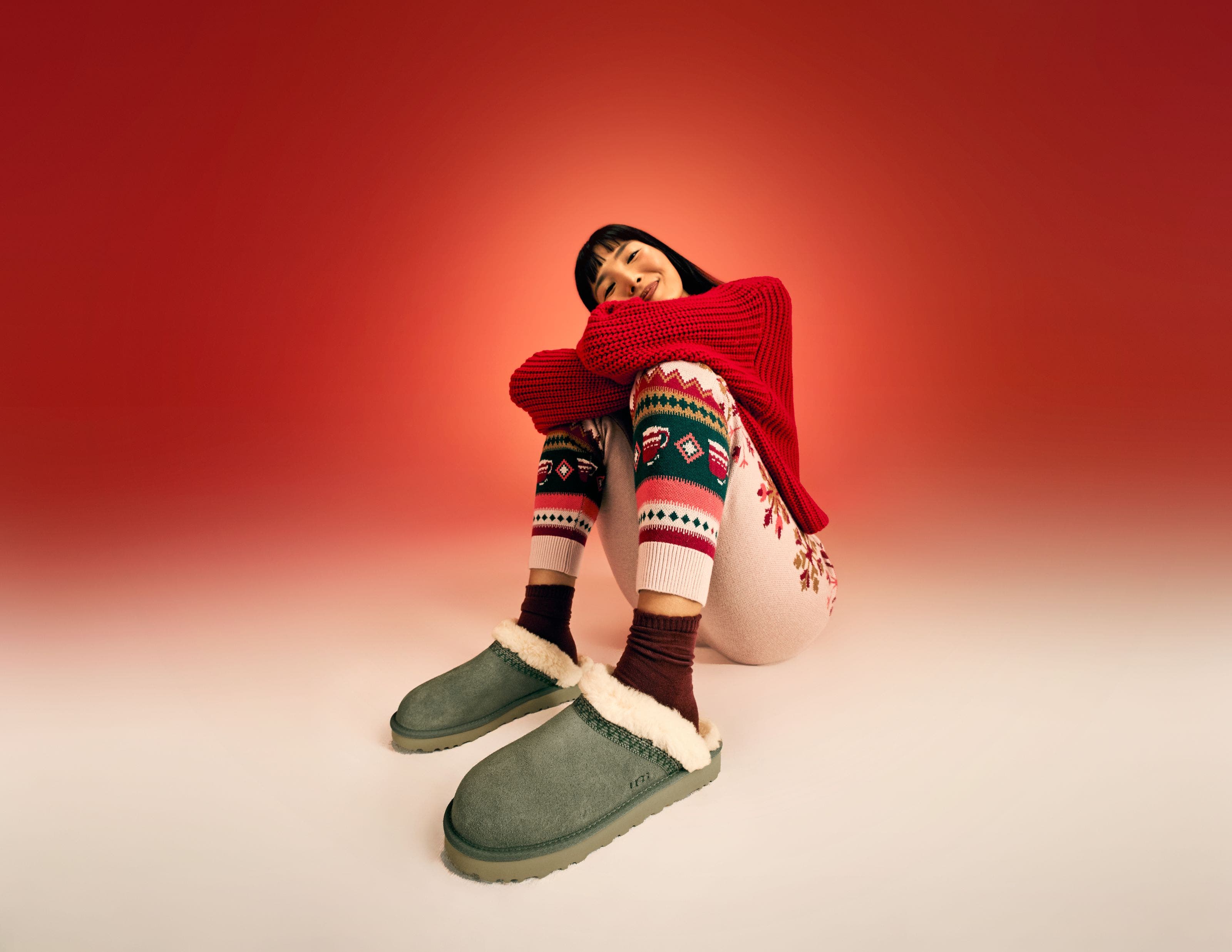 A woman wearing a red sweater, knit pants and UGG slippers. 