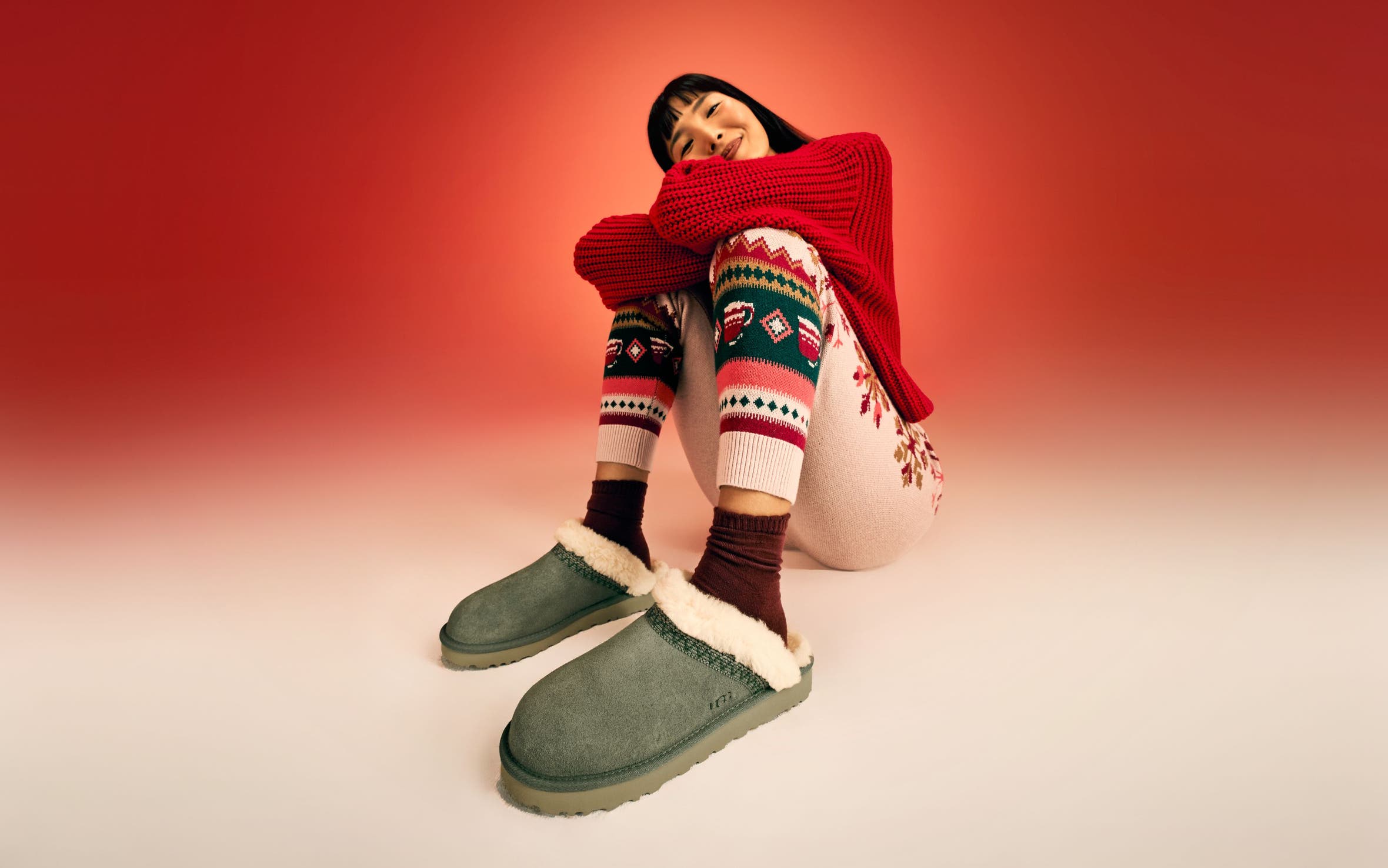 A woman wearing a red sweater, knit pants and UGG slippers.
