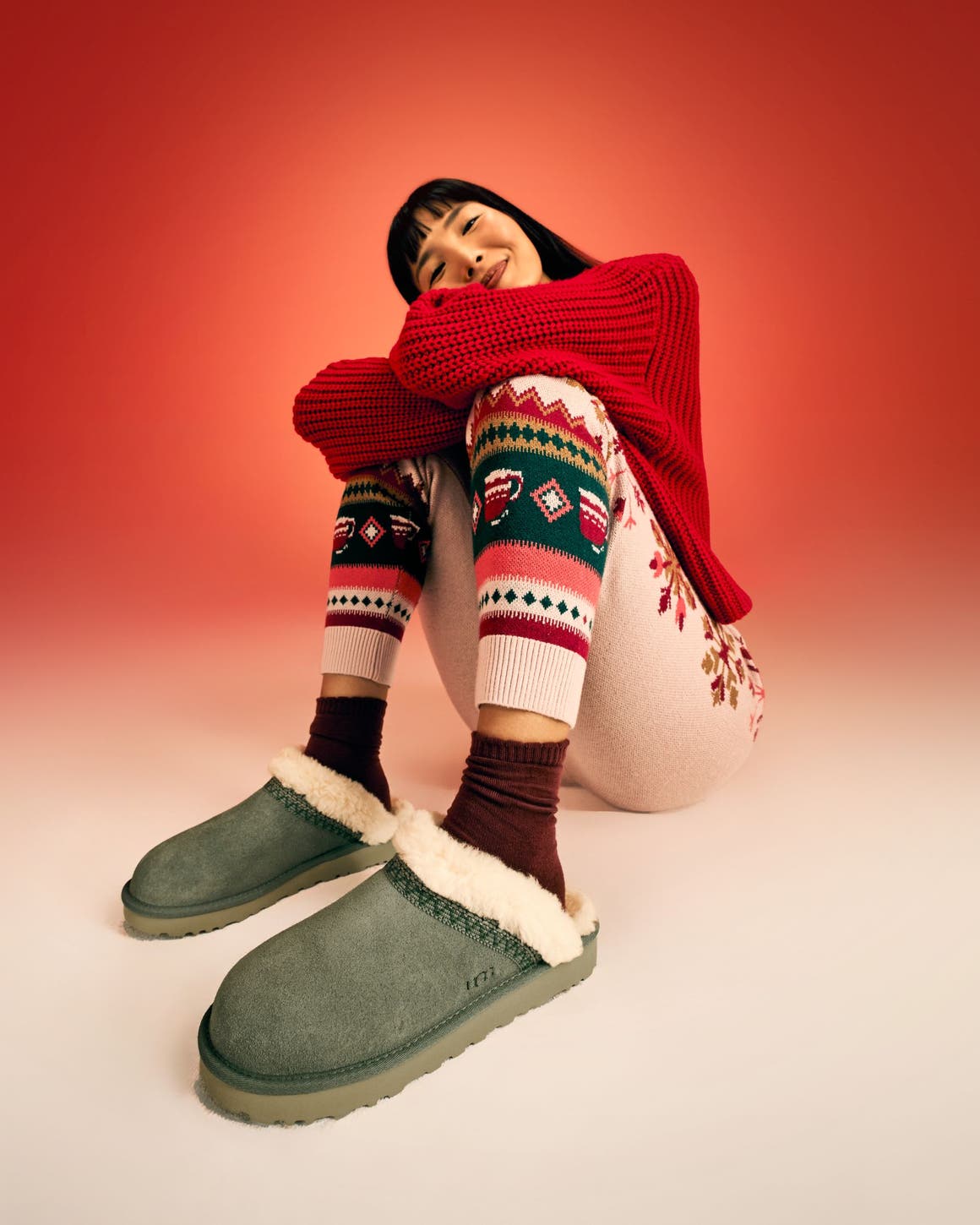 A woman wearing a red sweater, knit pants and UGG slippers.