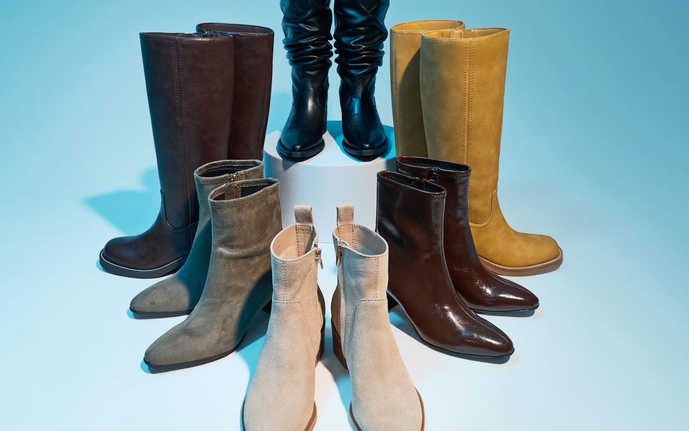 Seven pairs of women's boots arranged in a circle in various styles and colors against a teal blue background.