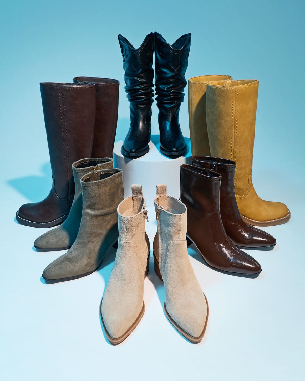 Seven pairs of women's boots arranged in a circle in various styles and colors against a teal blue background.