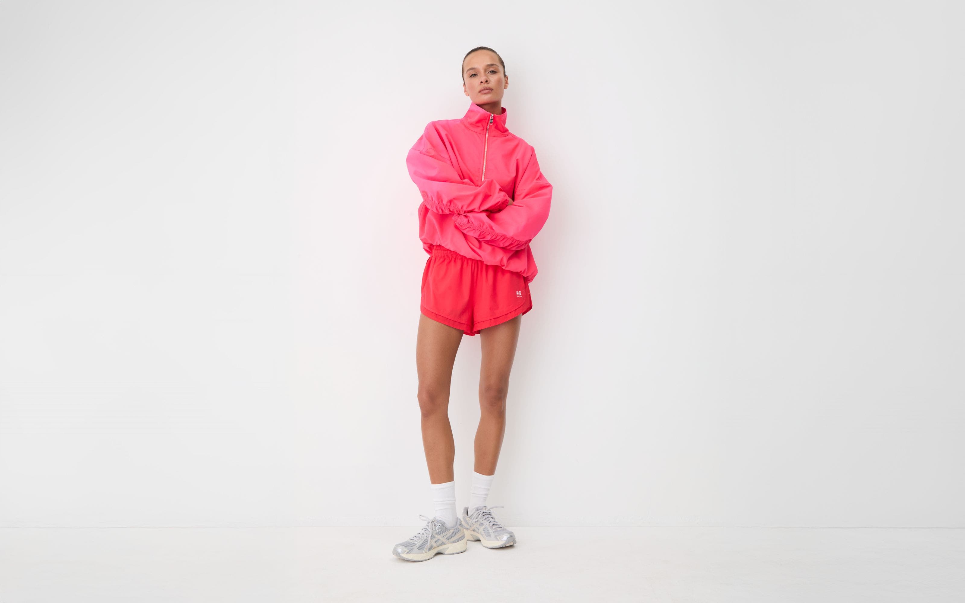 Woman in a pink half-zip pullover and pink runner shorts with white crew socks and silver sneakers.