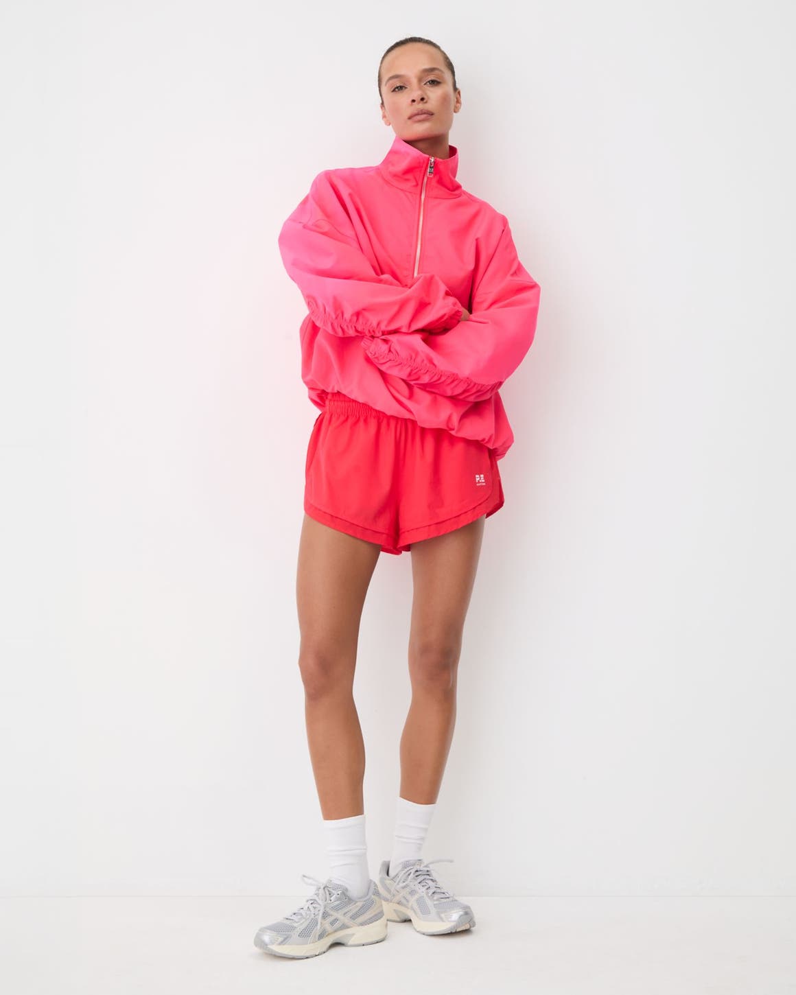Woman in a pink half-zip pullover and pink runner shorts with white crew socks and silver sneakers.