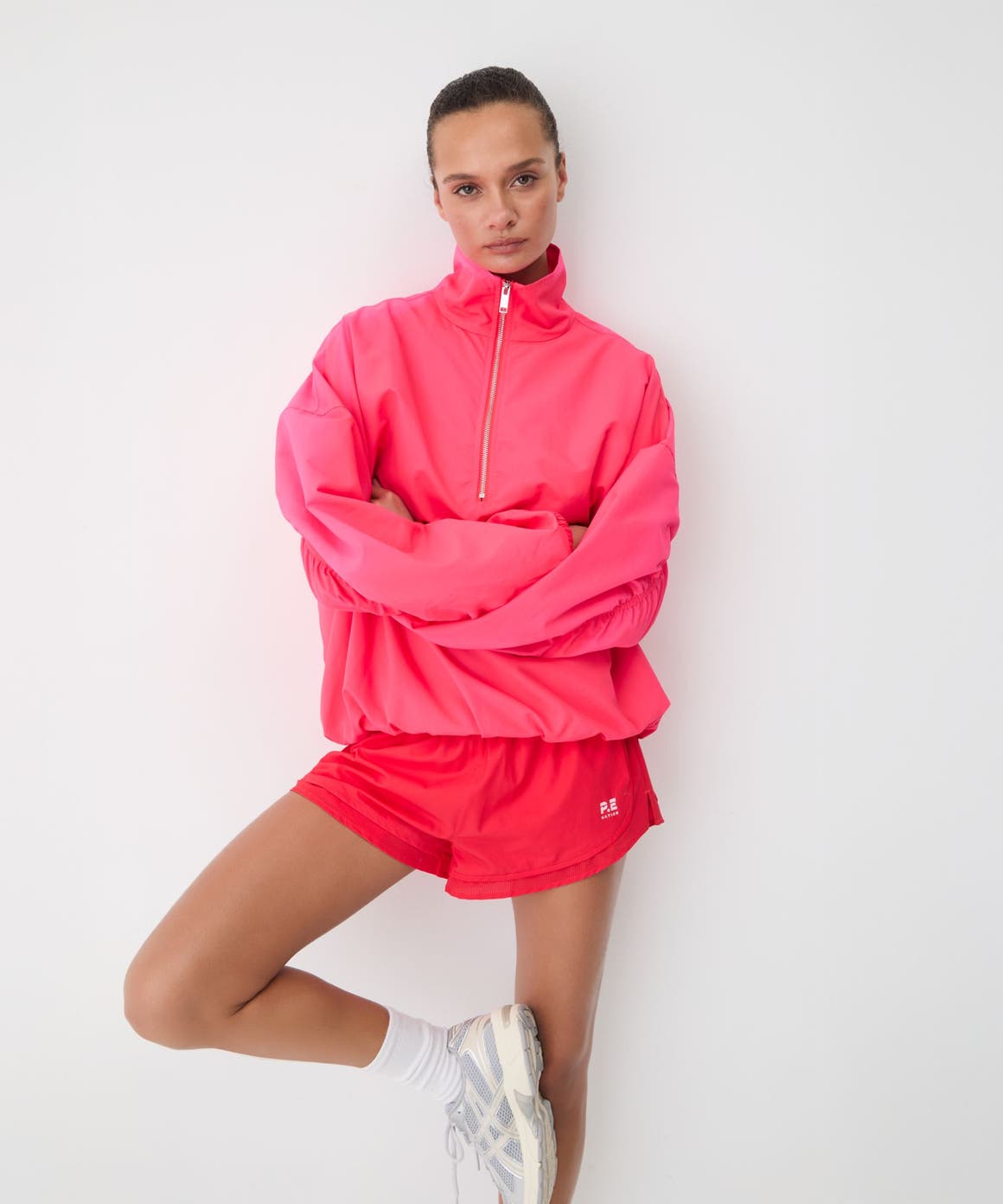 Woman in a pink half-zip pullover and pink runner shorts with white crew socks and silver sneakers.