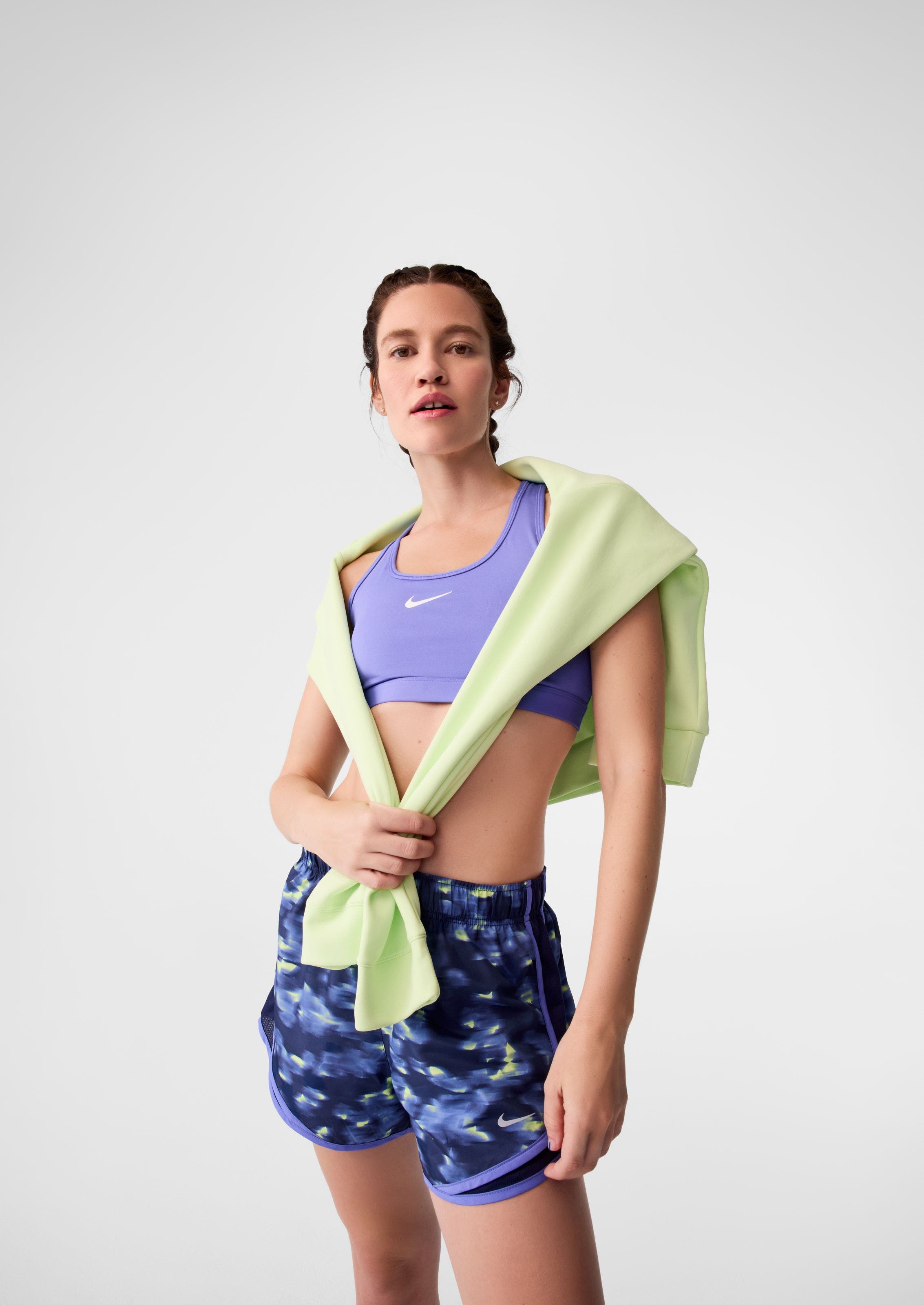 A woman wearing a purple sports bra, printed shorts and a green sweatshirt.