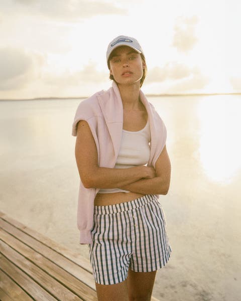 Woman wearing a baseball cap, white tank and striped shorts with a pink sweatshirt around her shoulders.