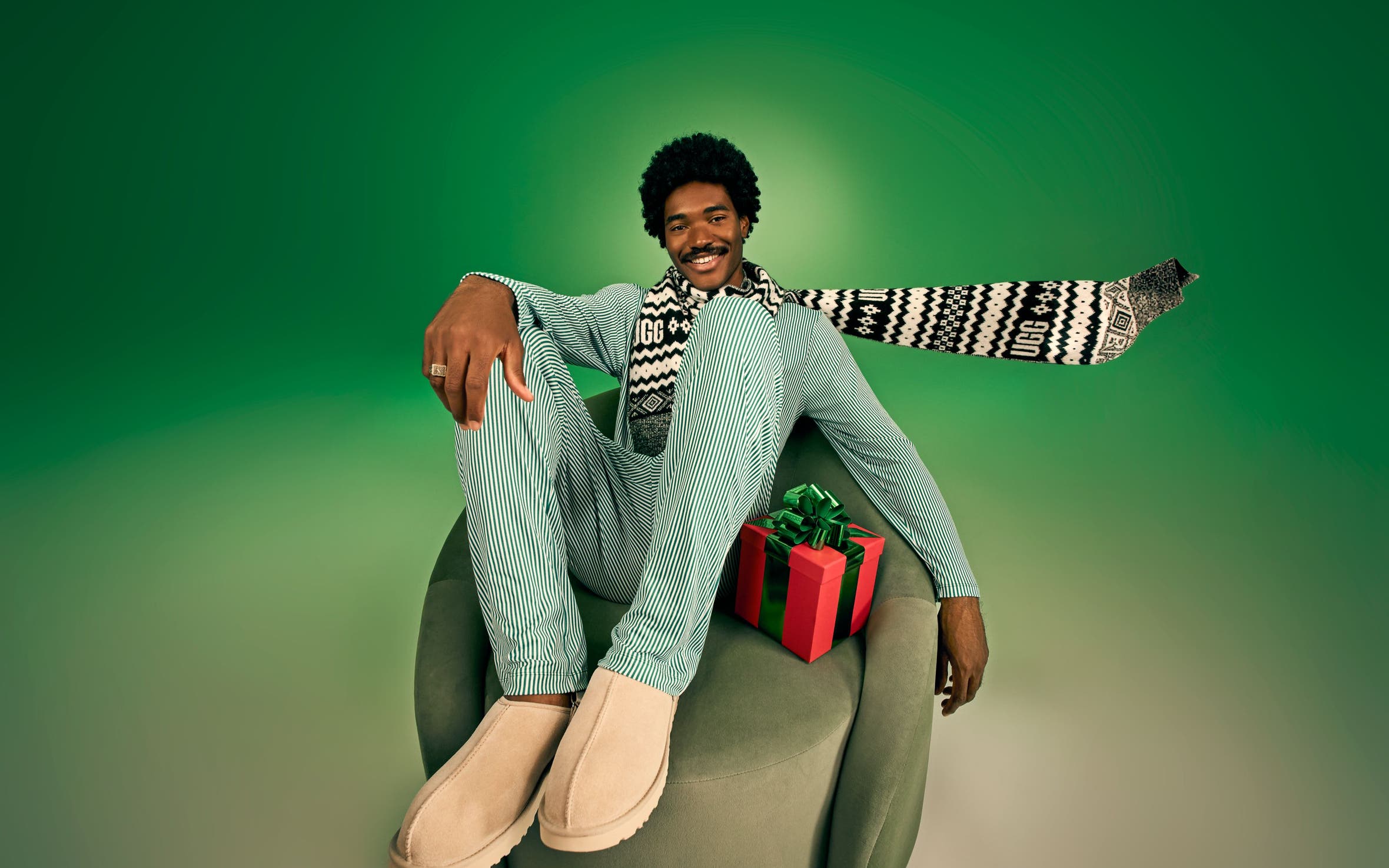 Male model wearing a green striped matching set, UGG patterned scarf, and tan UGG slippers seated in a green velvet chair with a holiday gift.