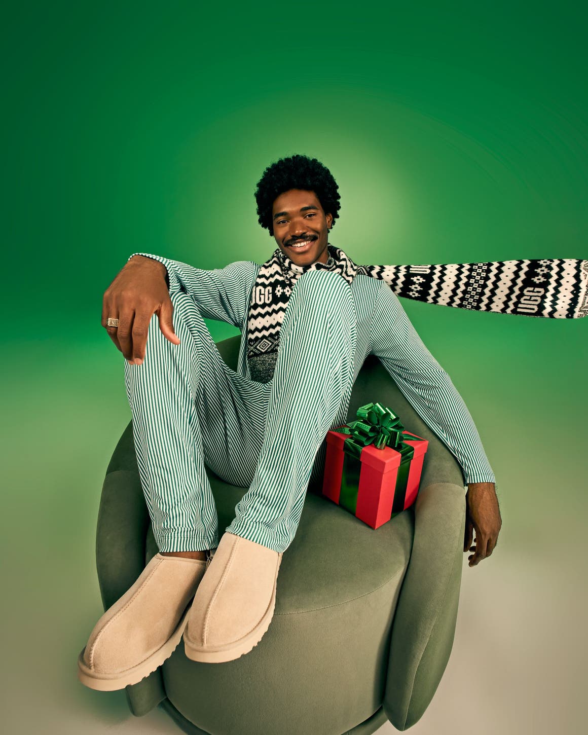 Male model wearing a green striped matching set, UGG patterned scarf, and tan UGG slippers seated in a green velvet chair with a holiday gift.