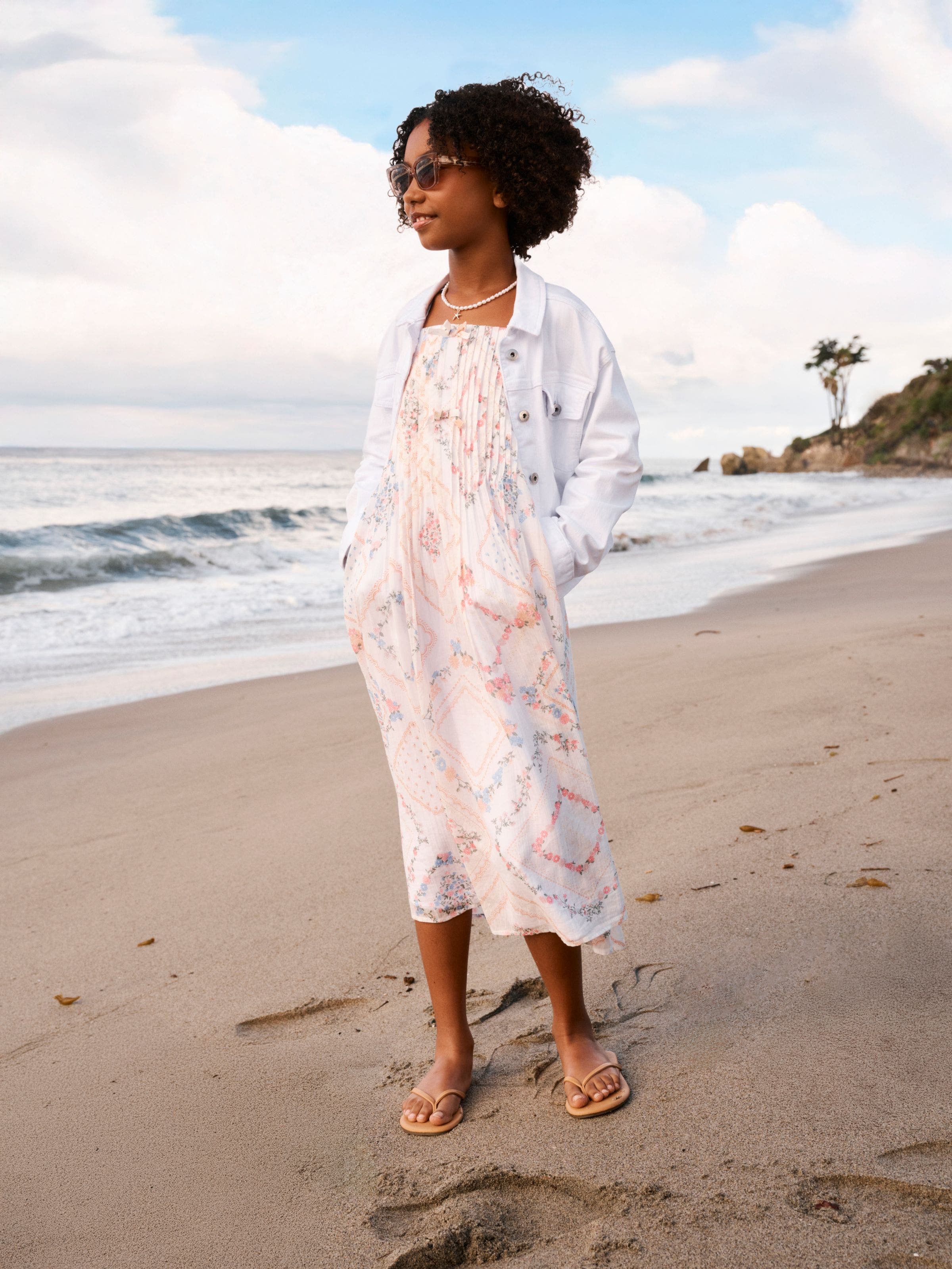 A tween girl wearing a sundress and a white denim jacket.