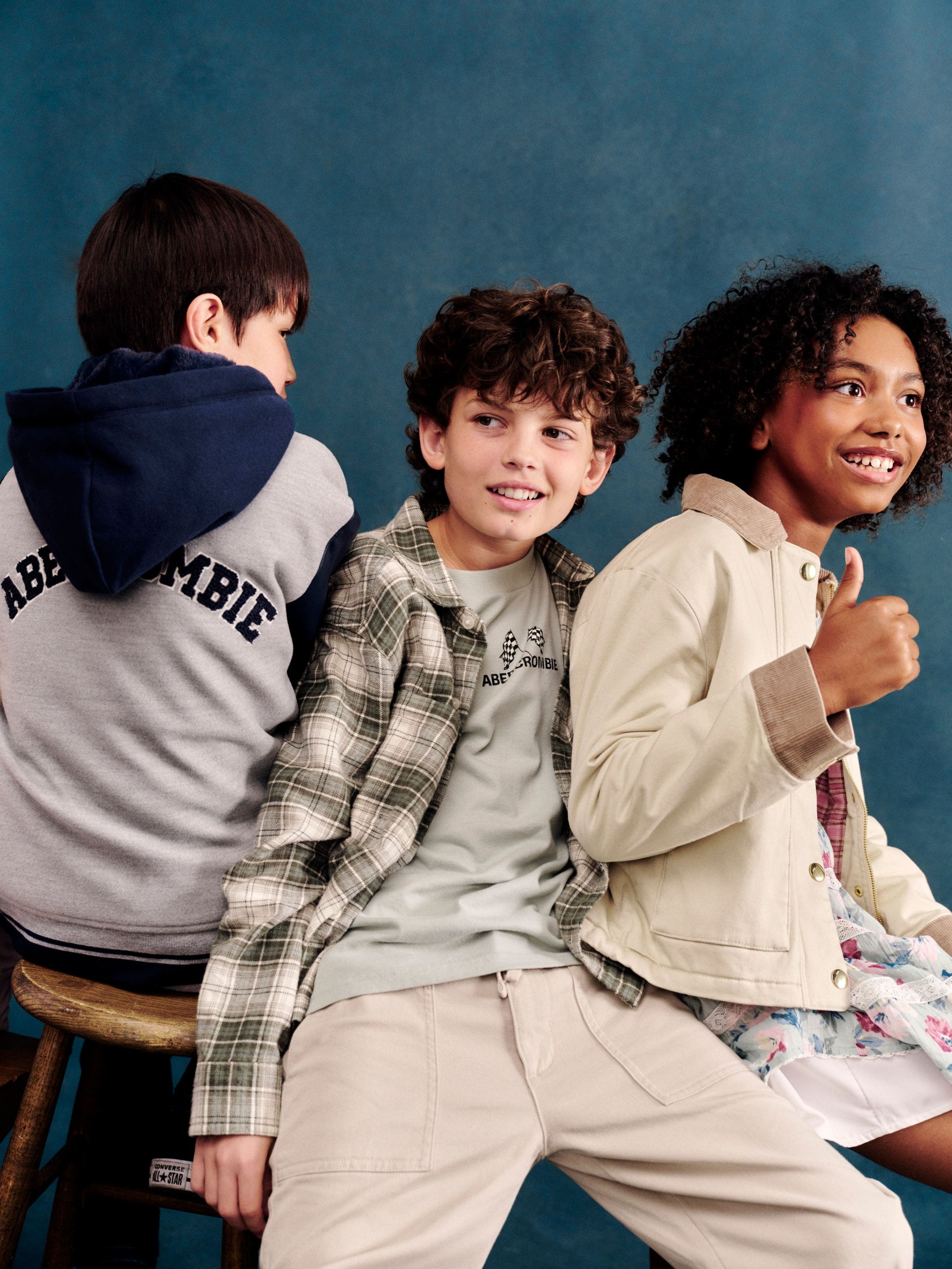 Three kids sitting together, wearing a gray logo hoodie, a plaid shirt layered over a graphic tee with khaki pants, and a tan utility jacket over a floral dress.