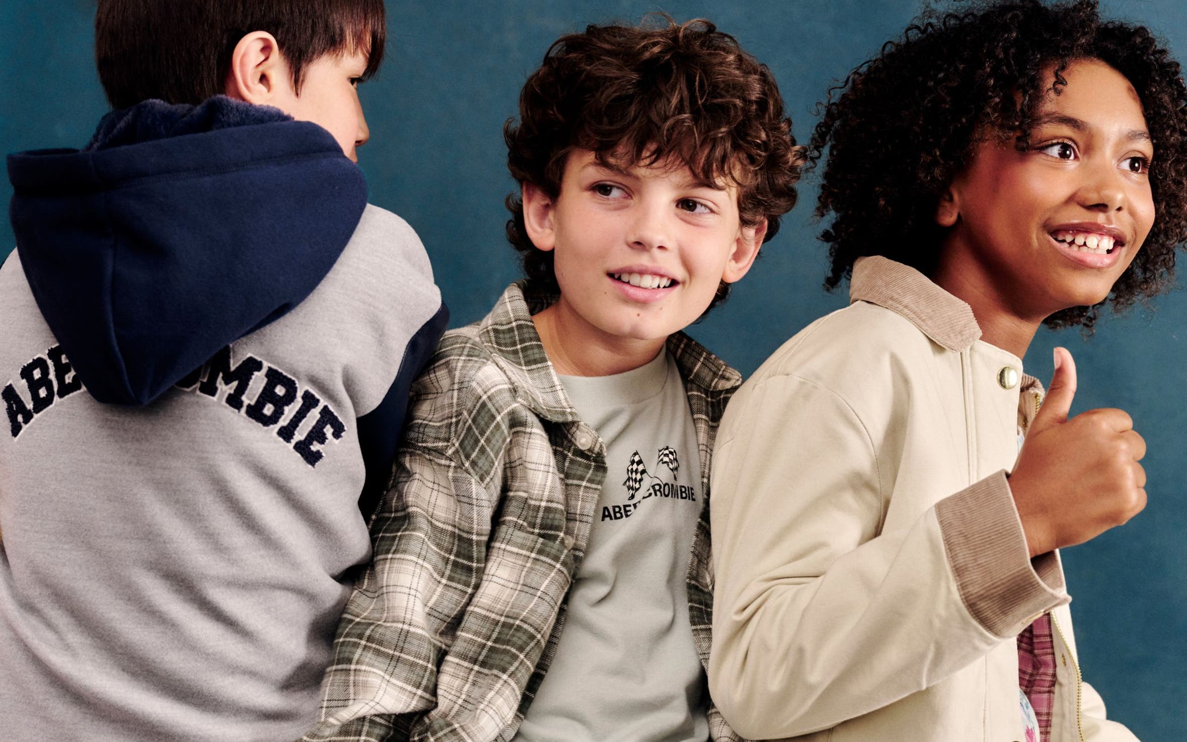 Three kids sitting together, wearing a gray logo hoodie, a plaid shirt layered over a graphic tee with khaki pants, and a tan utility jacket over a floral dress.