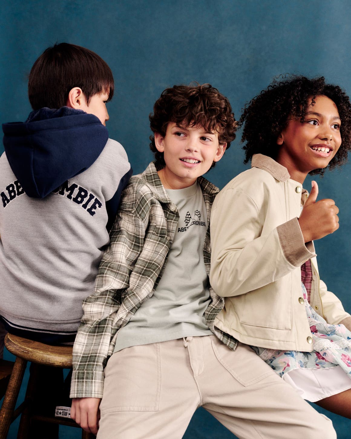 Three kids sitting together, wearing a gray logo hoodie, a plaid shirt layered over a graphic tee with khaki pants, and a tan utility jacket over a floral dress.