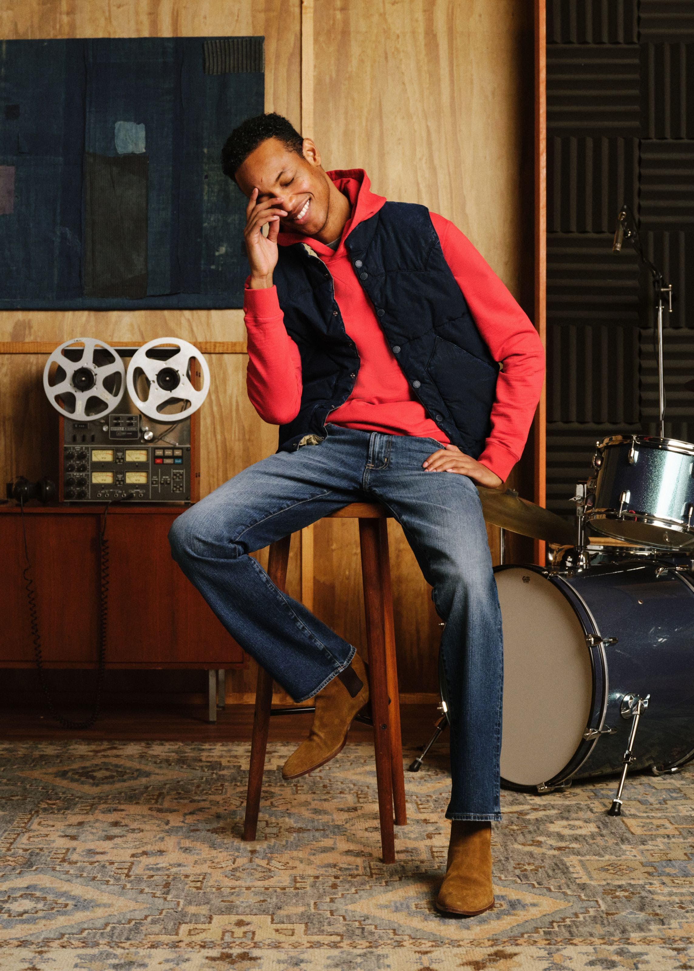 A man sitting down wearing ag denim, sweater, and vest.