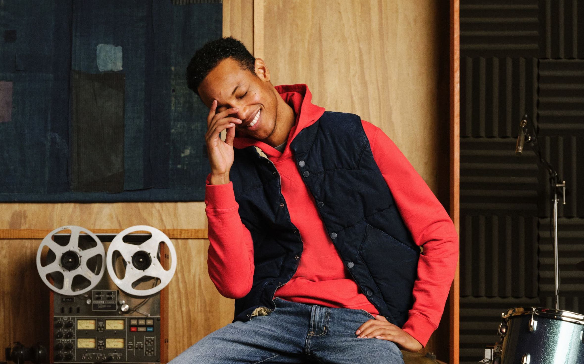 A man sitting down wearing ag denim, sweater, and vest.
