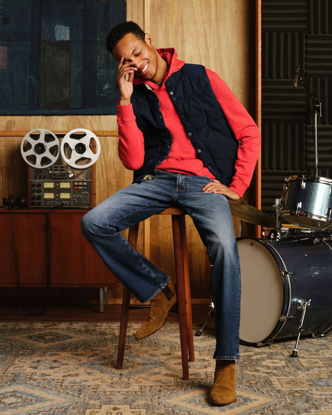 A man sitting down wearing ag denim, sweater, and vest.