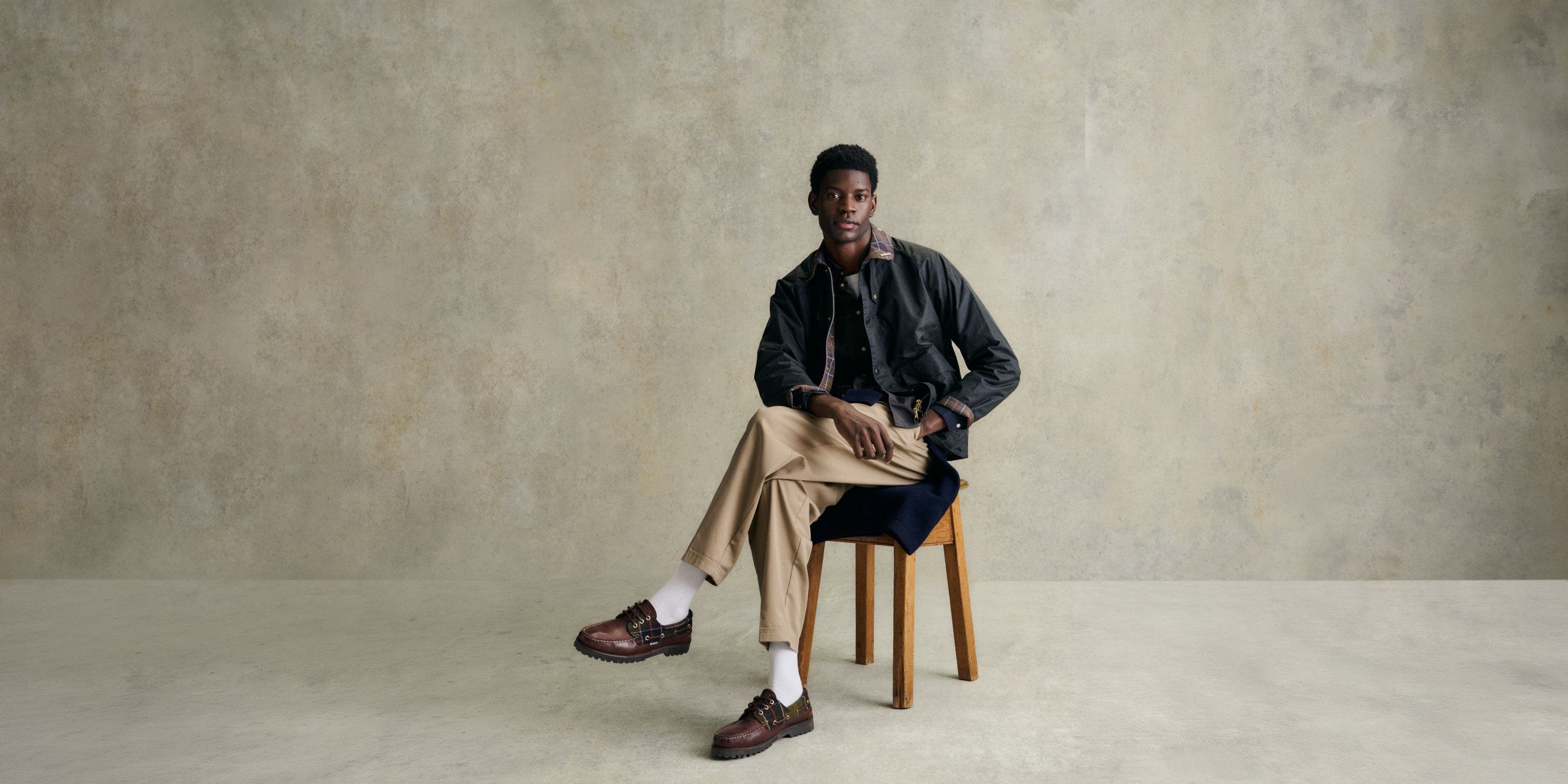 A man in Barbour Jacket, tan pants, and brown shoes sitting down.