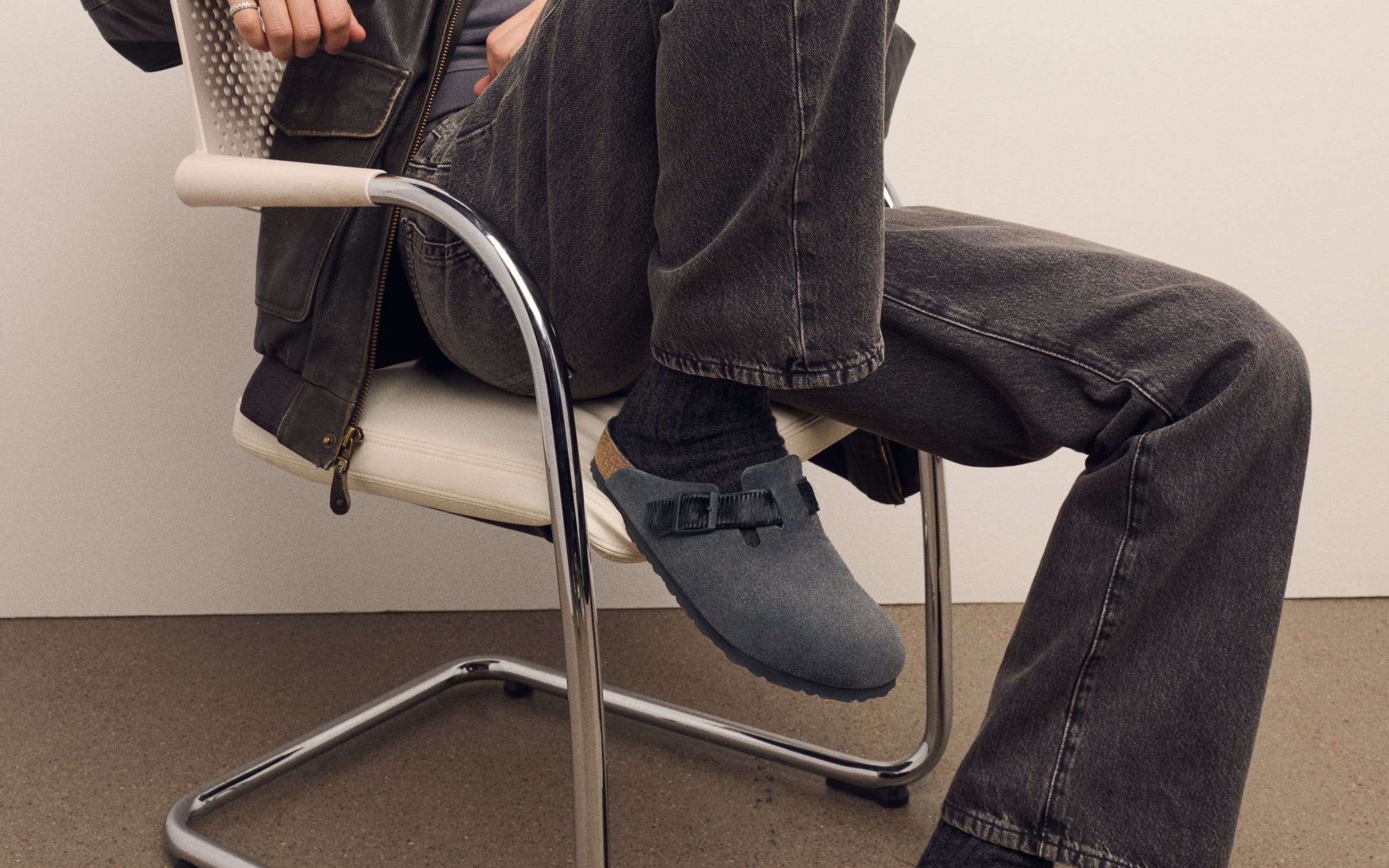 Man seated wearing Birkenstock Boston clogs in grey suede styled with dark wash jeans and a zip jacket.