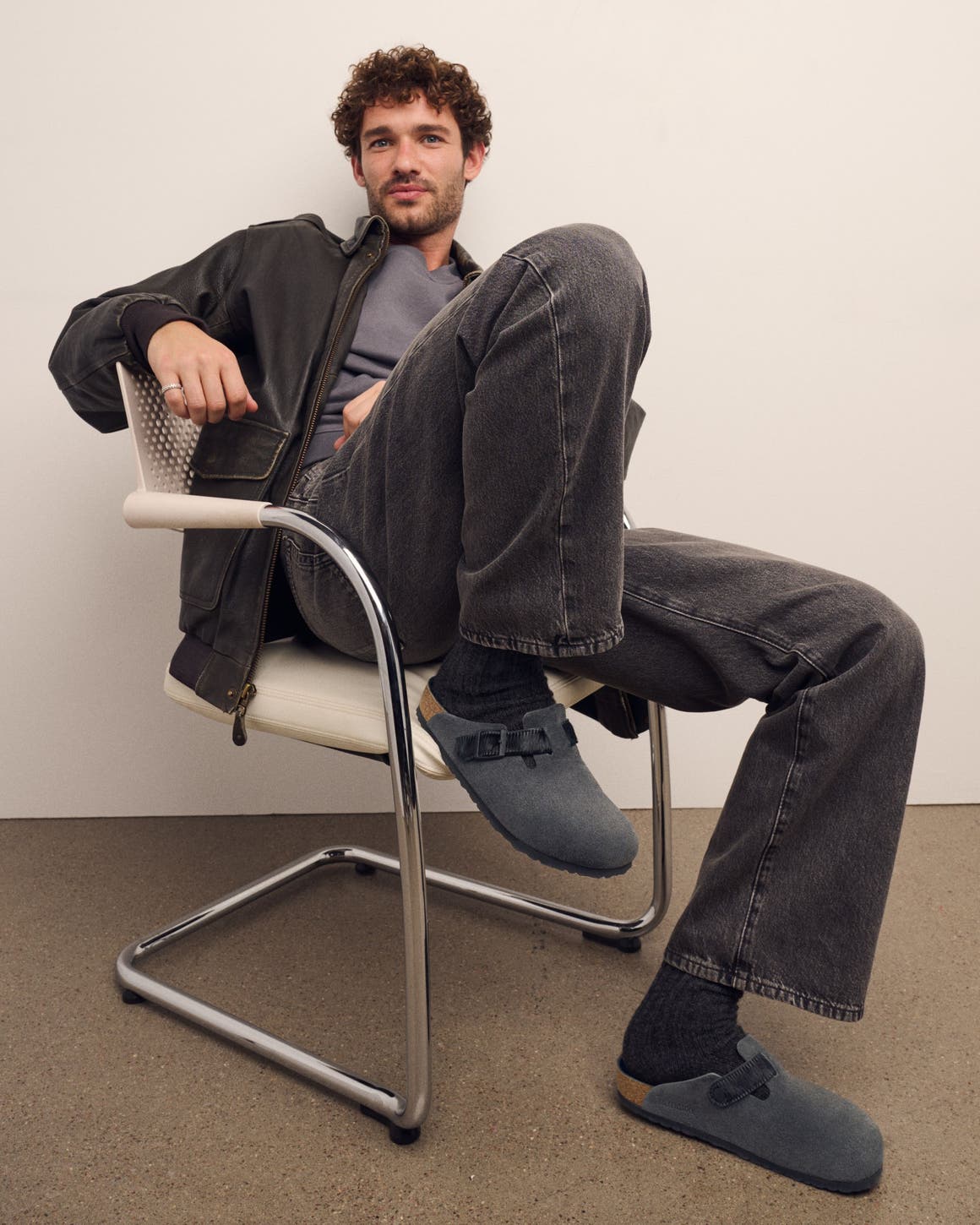 Man seated wearing Birkenstock Boston clogs in grey suede styled with dark wash jeans and a zip jacket.