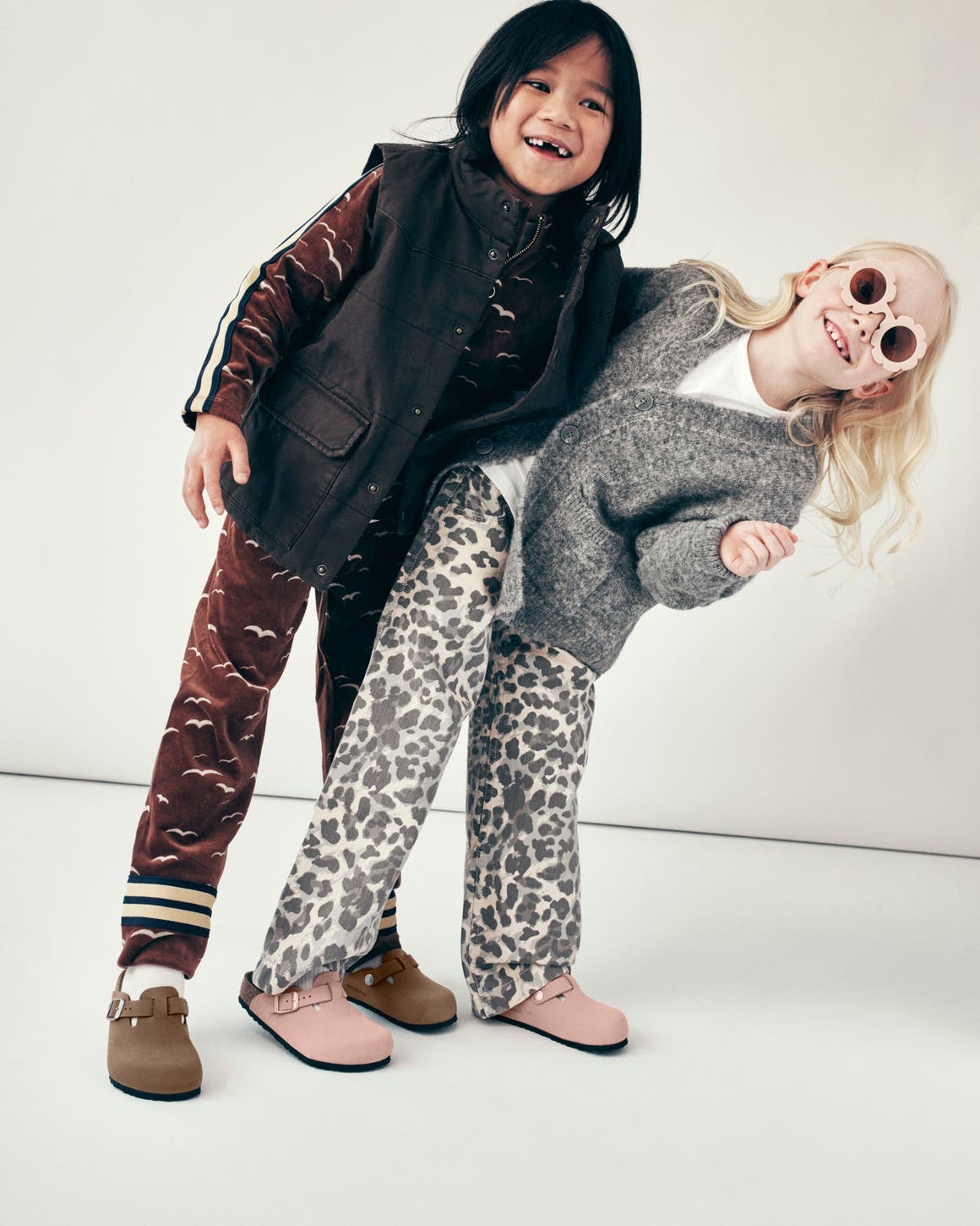 Two children wearing Birkenstock Boston clogs in tan suede and pink leather, styled in printed pants and layered outerwear.