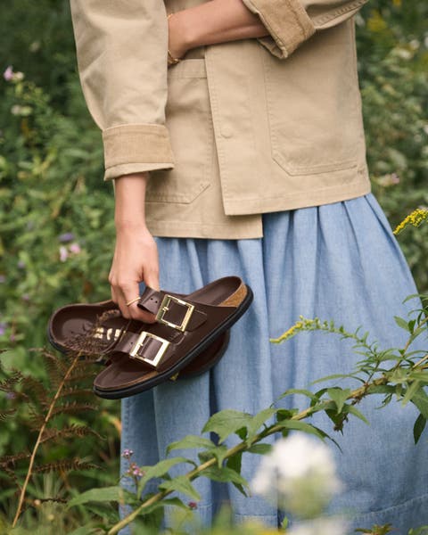 A woman standing in a field holding a pair of brown Birkenstock sandals.