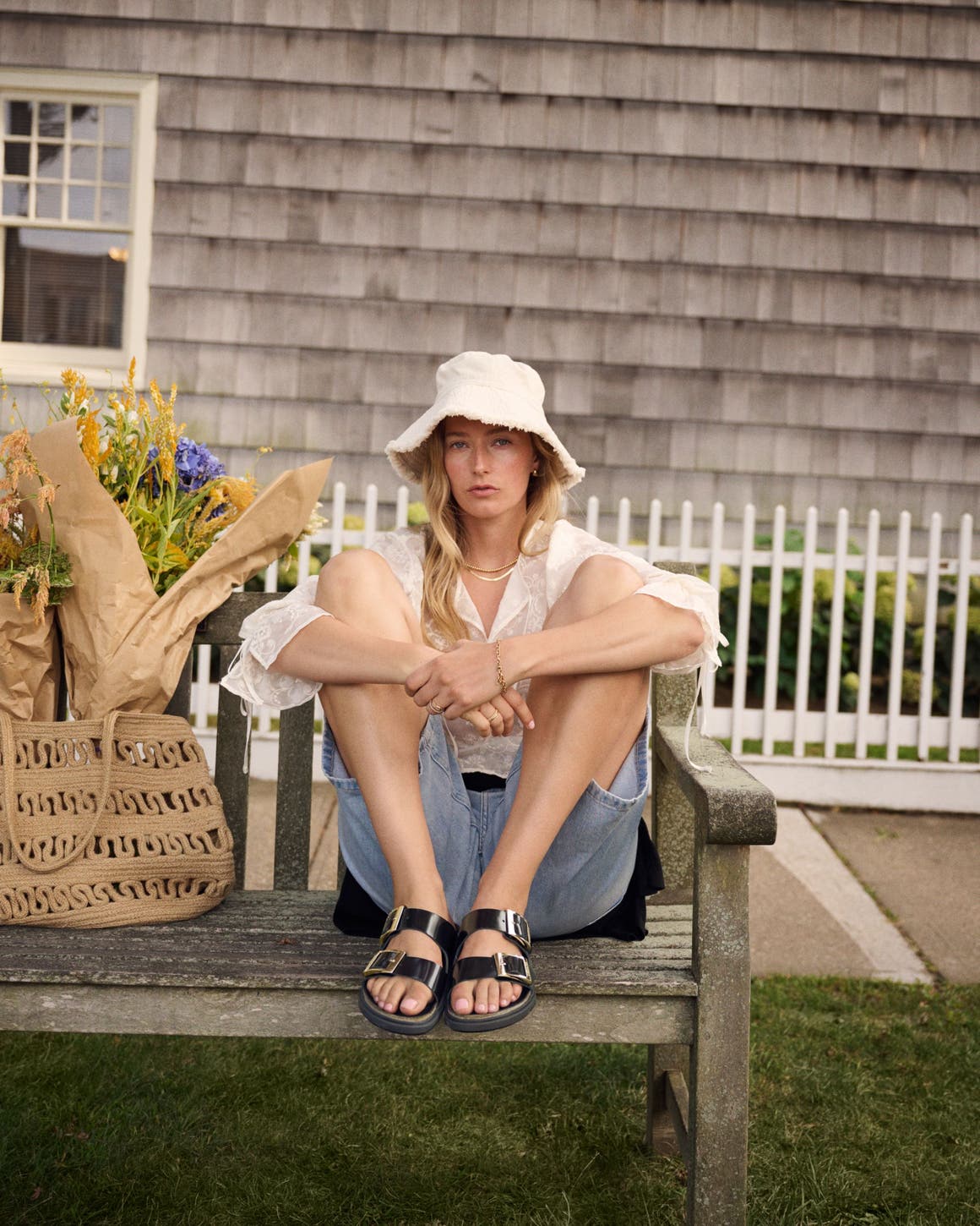 A woman on a weathered bench in light denim jeans, a white sun hat and black leather Birkenstocks sandals.