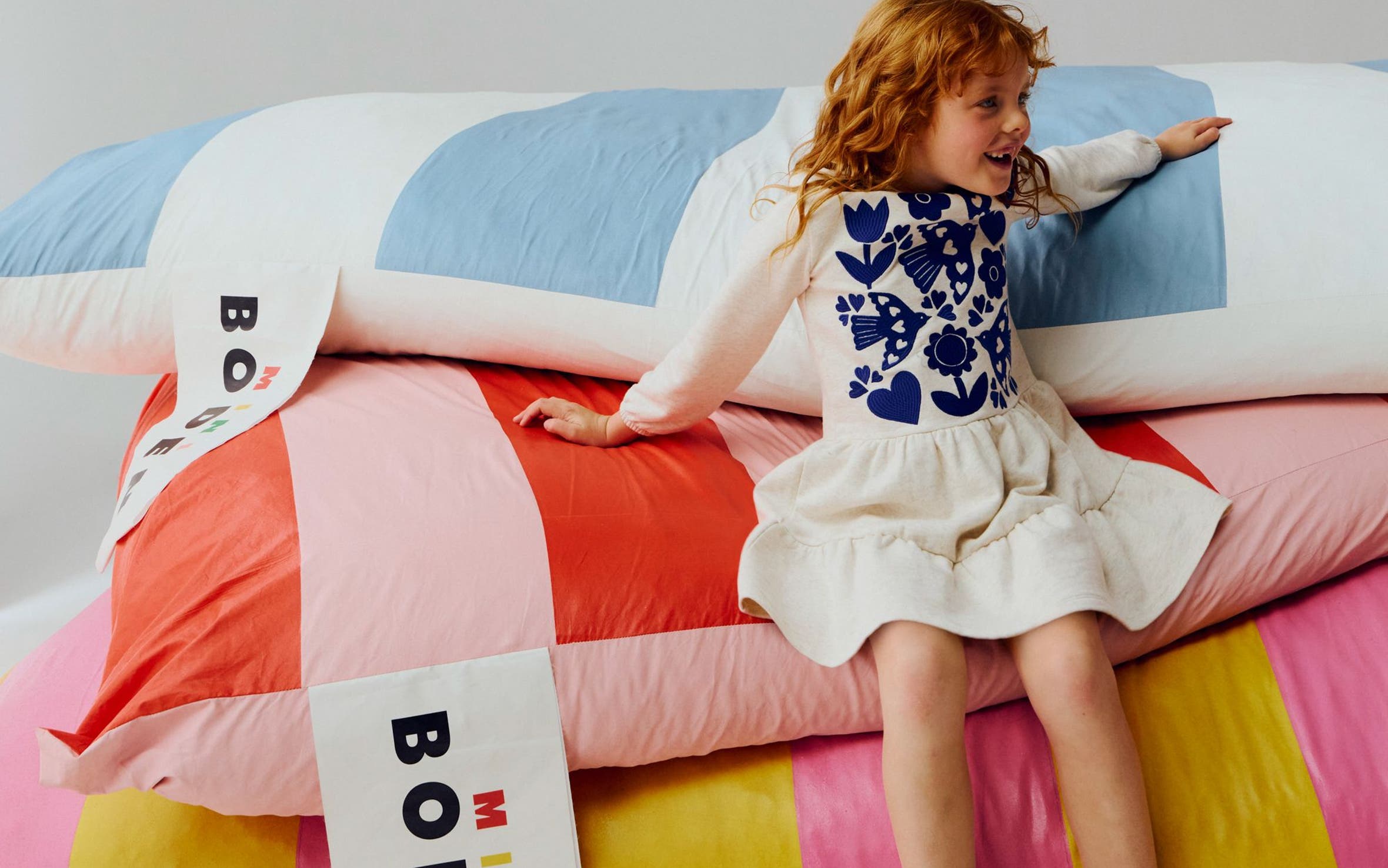 A girl wearing an embroidered dress sitting on striped Mini Boden cushions.
