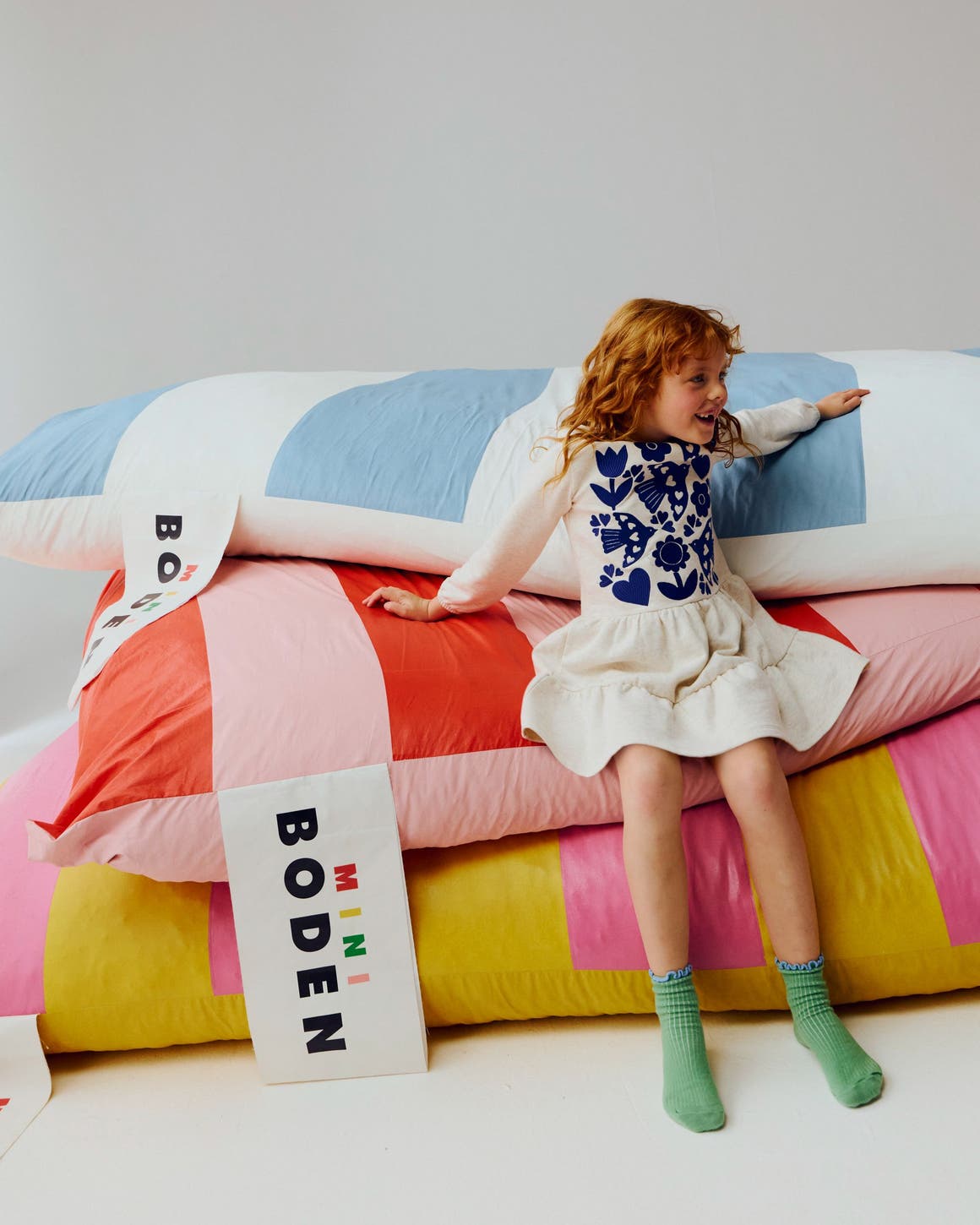 A girl wearing an embroidered dress sitting on striped Mini Boden cushions.