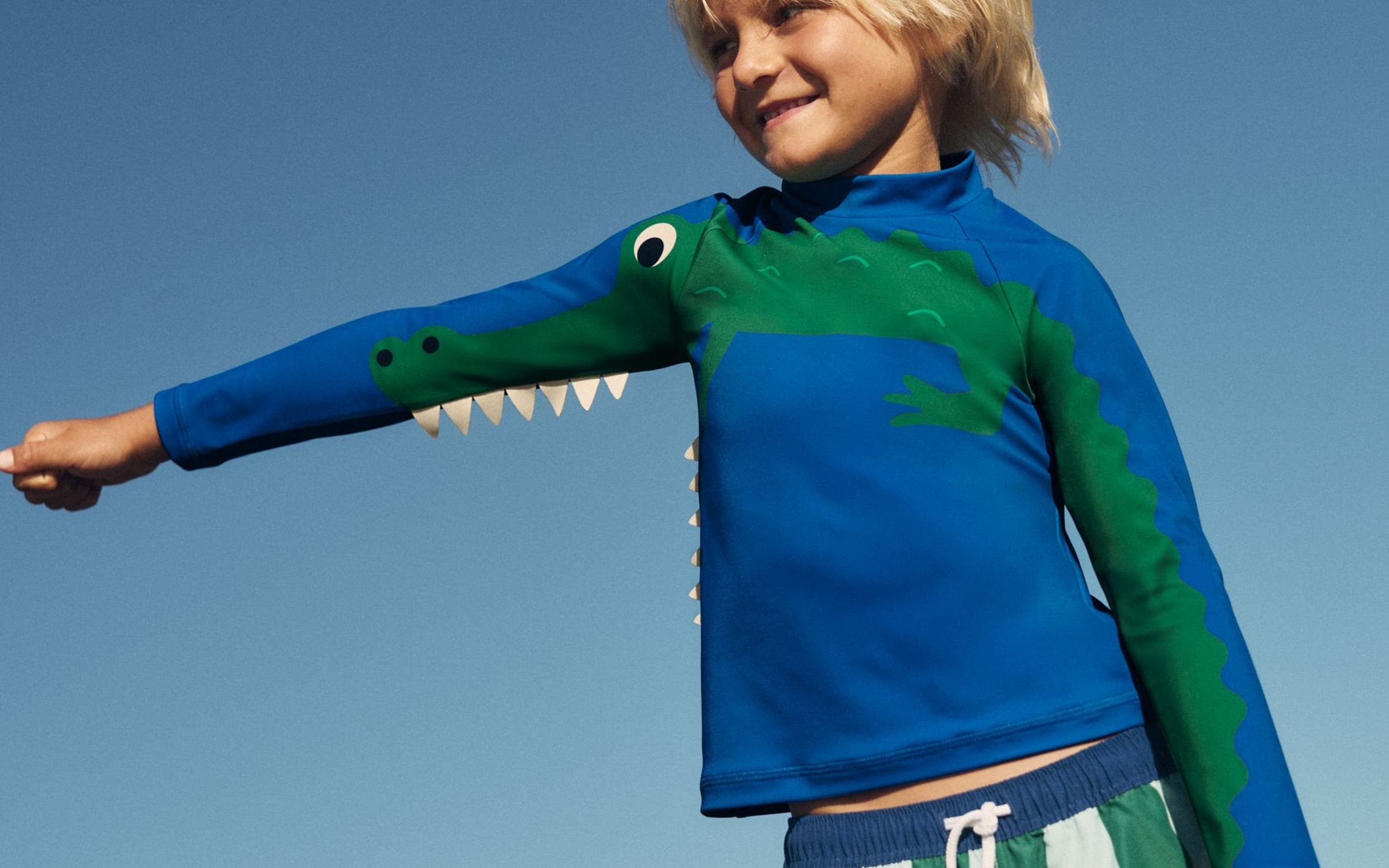 A boy wearing a Mini Boden rash guard and swim shorts.