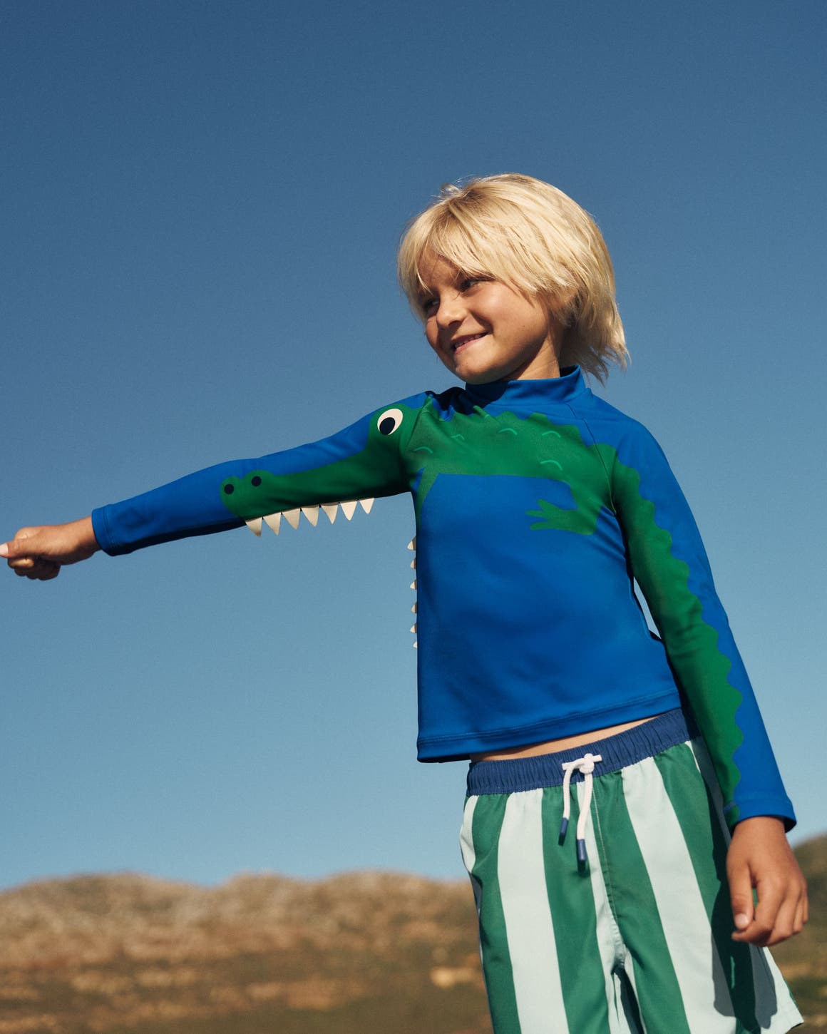 A boy wearing a Mini Boden rash guard and swim shorts.