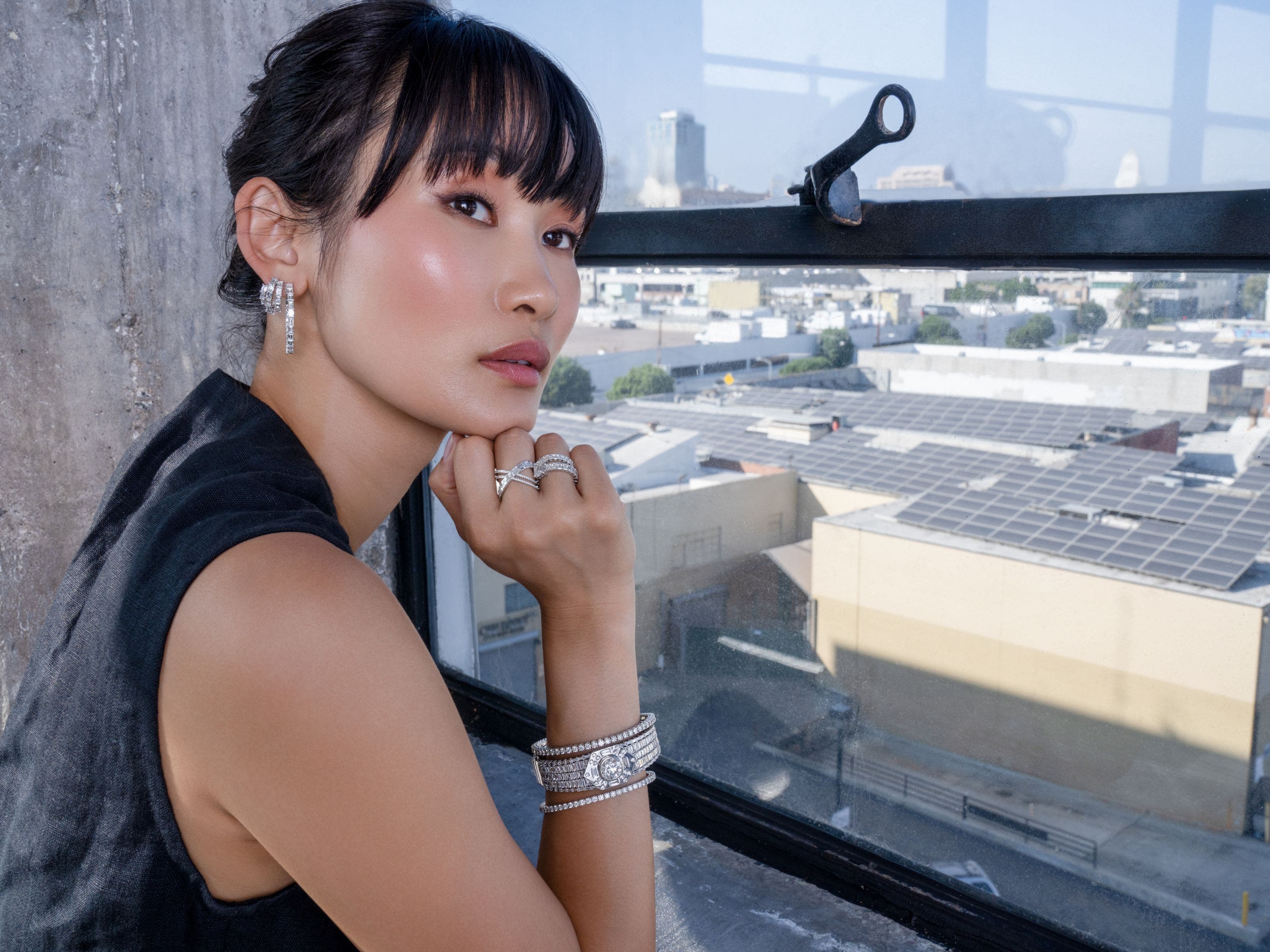 A woman leaning in front of a window with a city view, wearing diamond earrings, rings and bracelets.