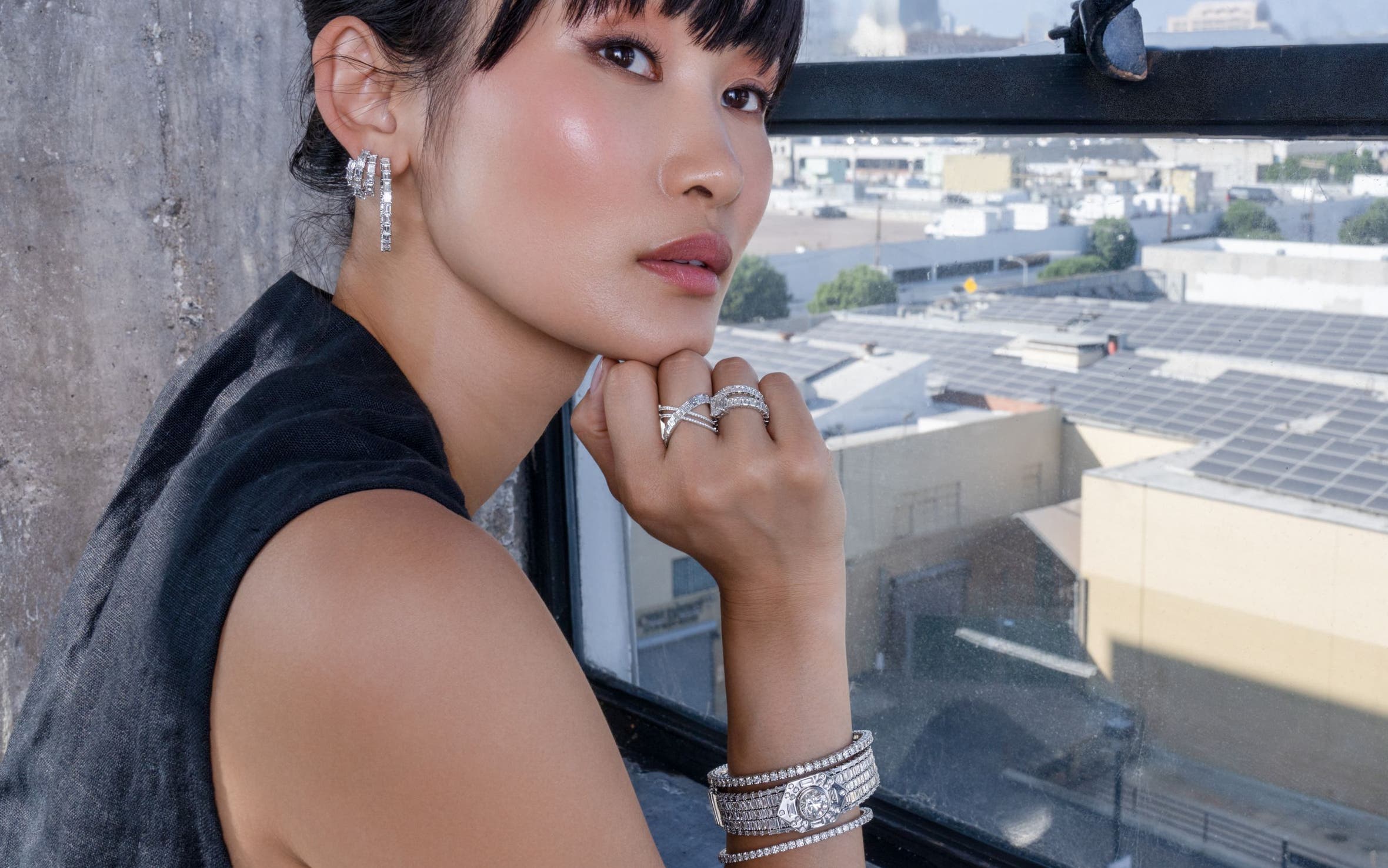 A woman leaning in front of a window with a city view, wearing diamond earrings, rings and bracelets.