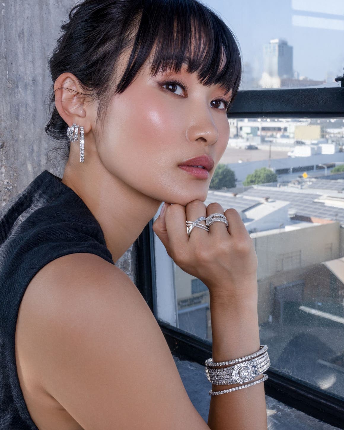 A woman leaning in front of a window with a city view, wearing diamond earrings, rings and bracelets.
